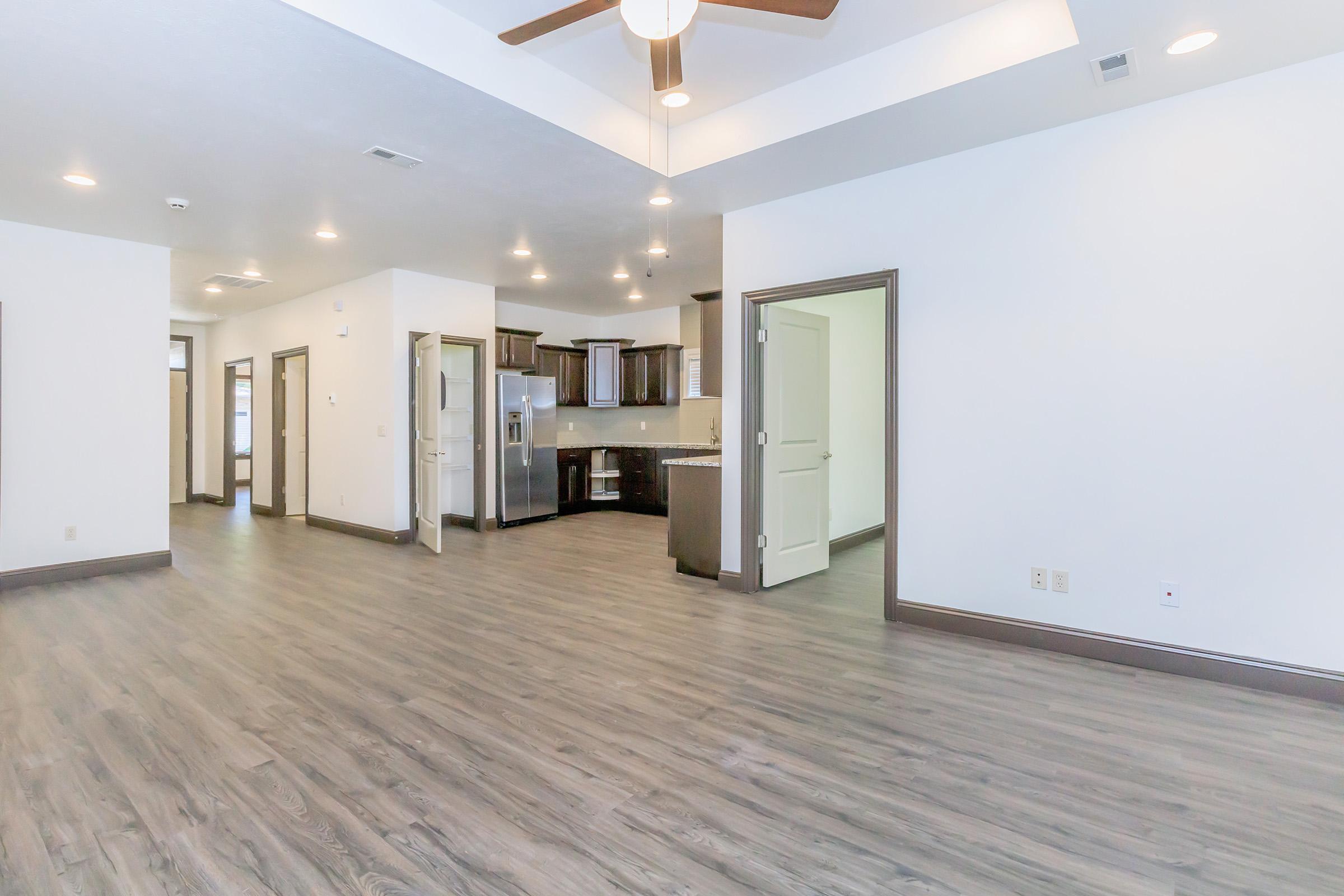 A spacious, modern interior featuring light gray walls and a wood-style floor. The open layout includes a kitchen with dark cabinetry and stainless steel appliances visible in the background. Ceiling fans and recessed lighting enhance the bright atmosphere, with multiple doorways leading to other areas.