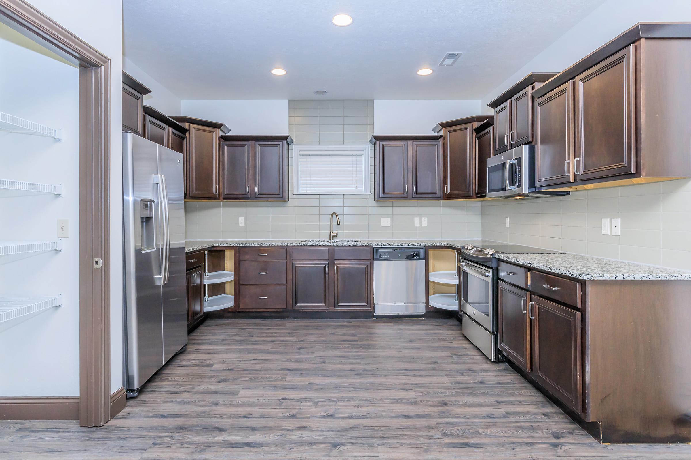 Modern kitchen featuring dark wood cabinets, stainless steel appliances, a granite countertop, and a spacious layout. Includes a dishwasher, refrigerator, and a double sink, with a pantry on the left. The flooring is a dark wood laminate, and the walls are a light neutral color, creating an inviting atmosphere.