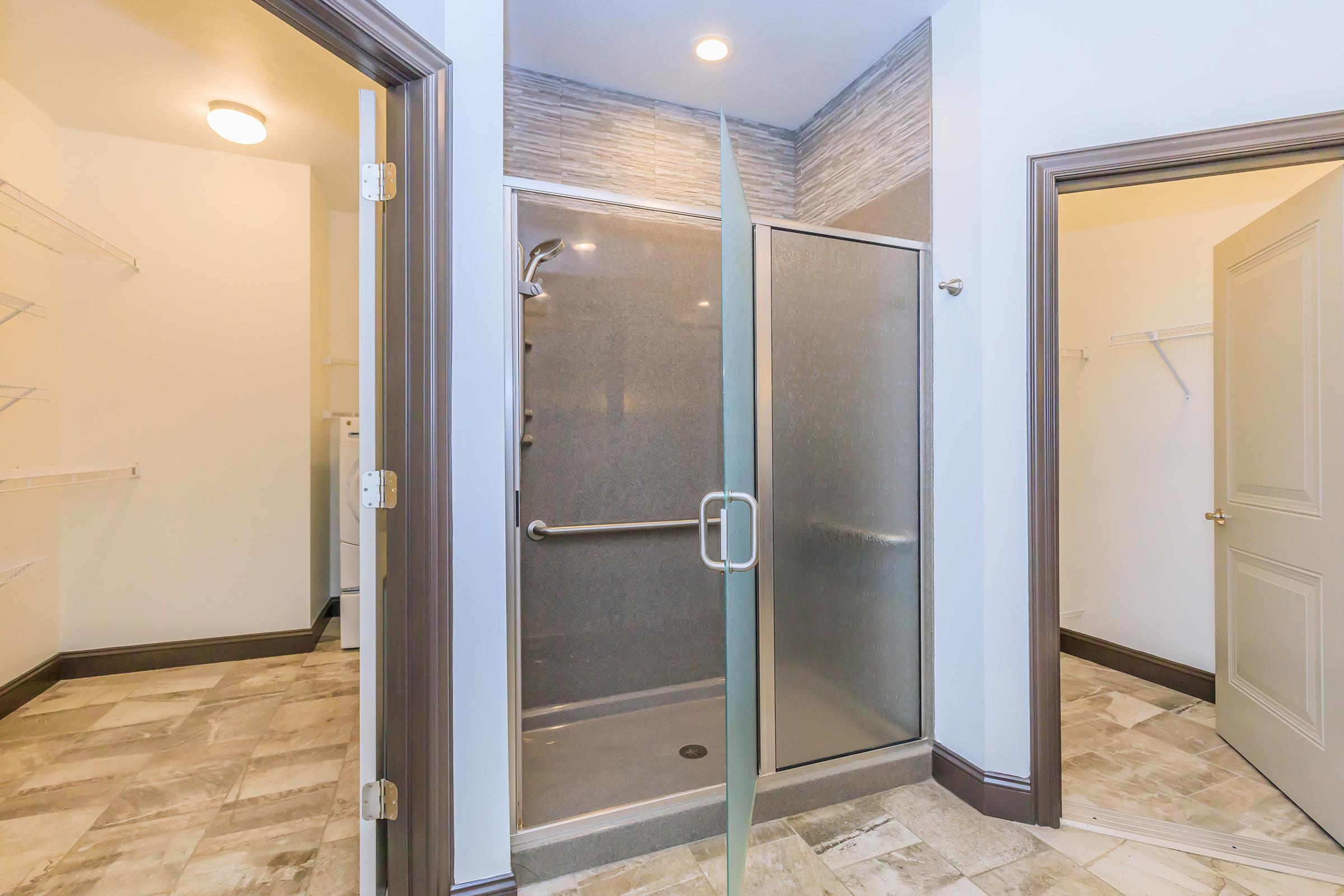 A modern bathroom featuring a glass-enclosed walk-in shower with a sliding door, neutral tiled flooring, and light-colored walls. There are shelves on one side and a door leading to another room, suggesting efficient use of space. Overall, the design is clean and contemporary.