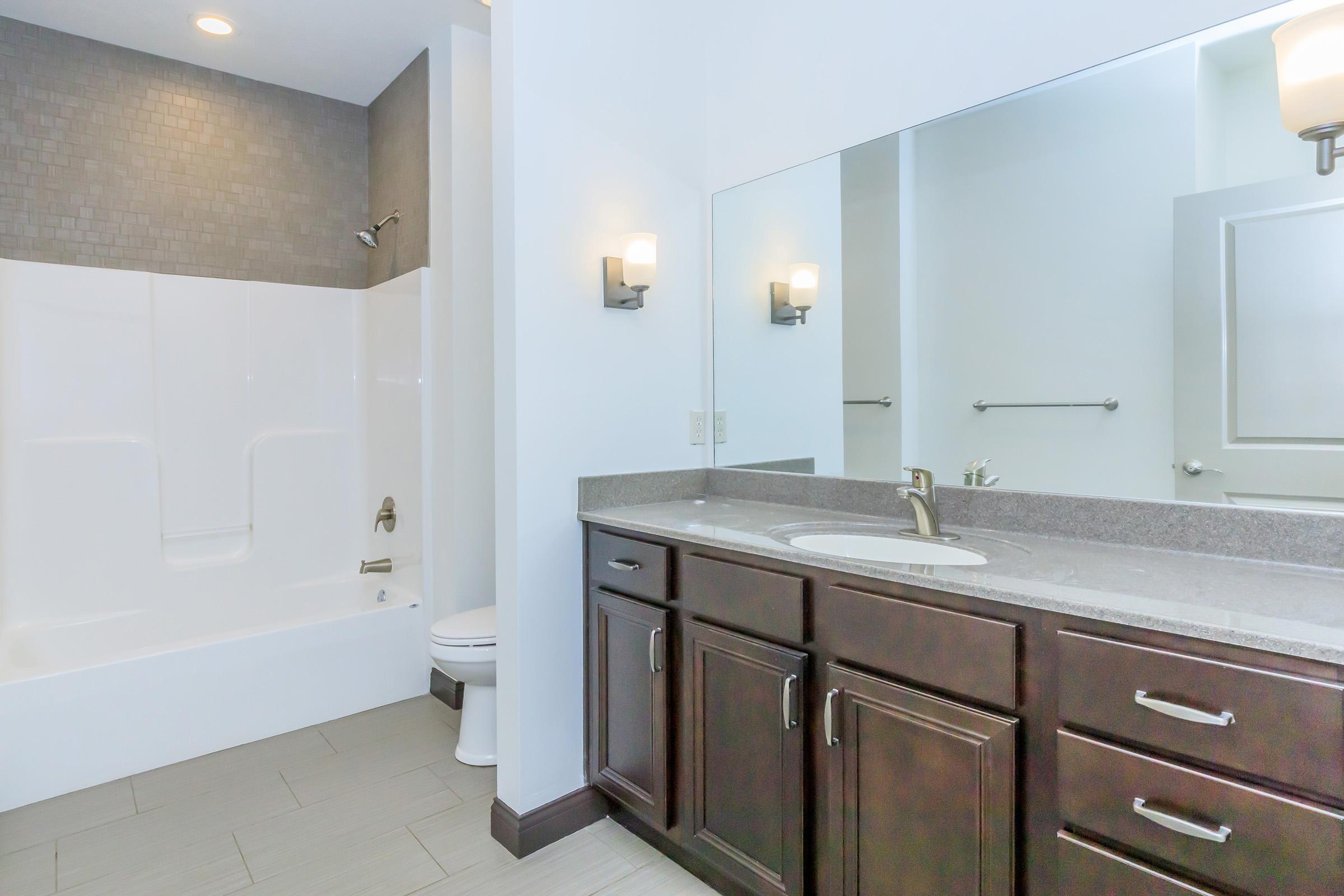 A modern bathroom featuring a white bathtub and shower combination, dark wood cabinetry with a granite countertop, a large mirror, and two wall-mounted light fixtures. Neutral-colored walls and tiled flooring enhance the contemporary aesthetic.