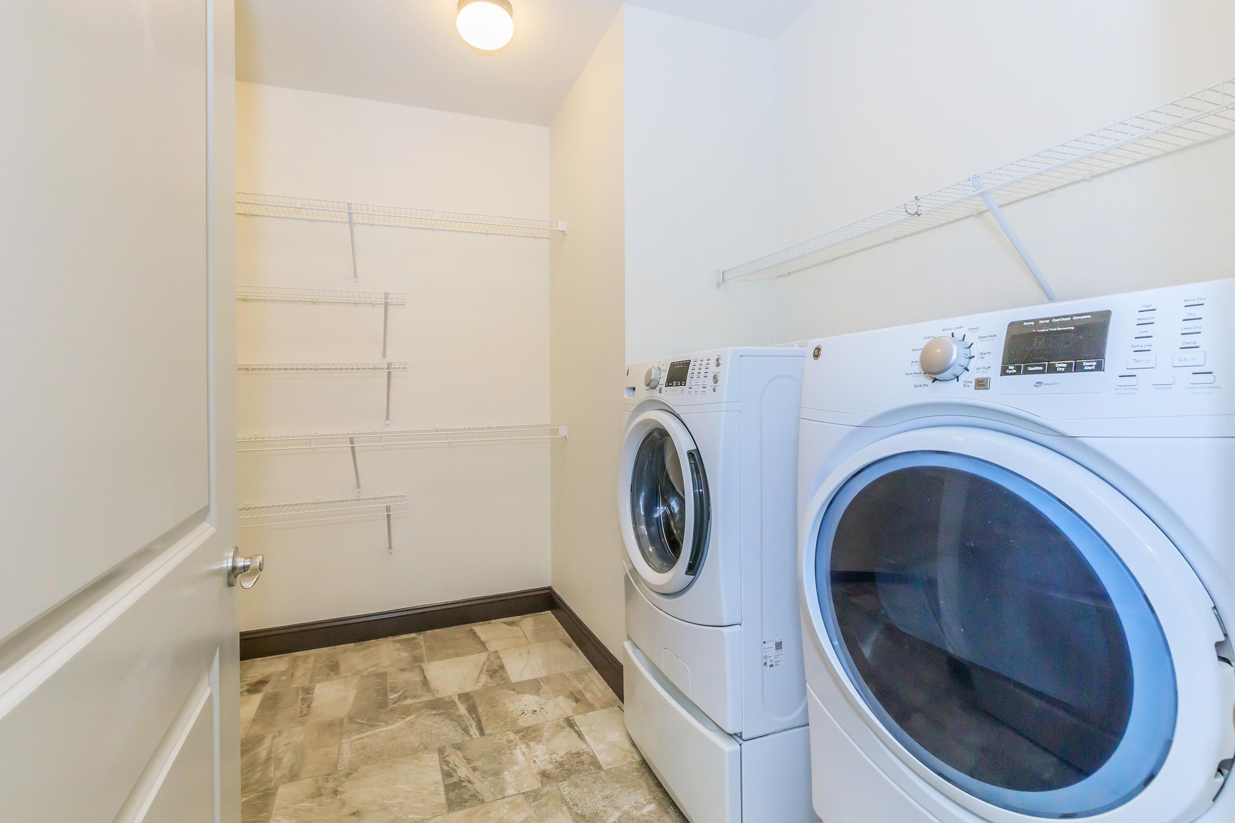 A bright and spacious laundry room featuring a white washer and dryer stacked on top of each other. The walls are painted light beige, and there's a small ceiling light. To the side, there's a set of metal wire shelves mounted on the wall, with plenty of open space for storage. The floor has a gray and white tile pattern.