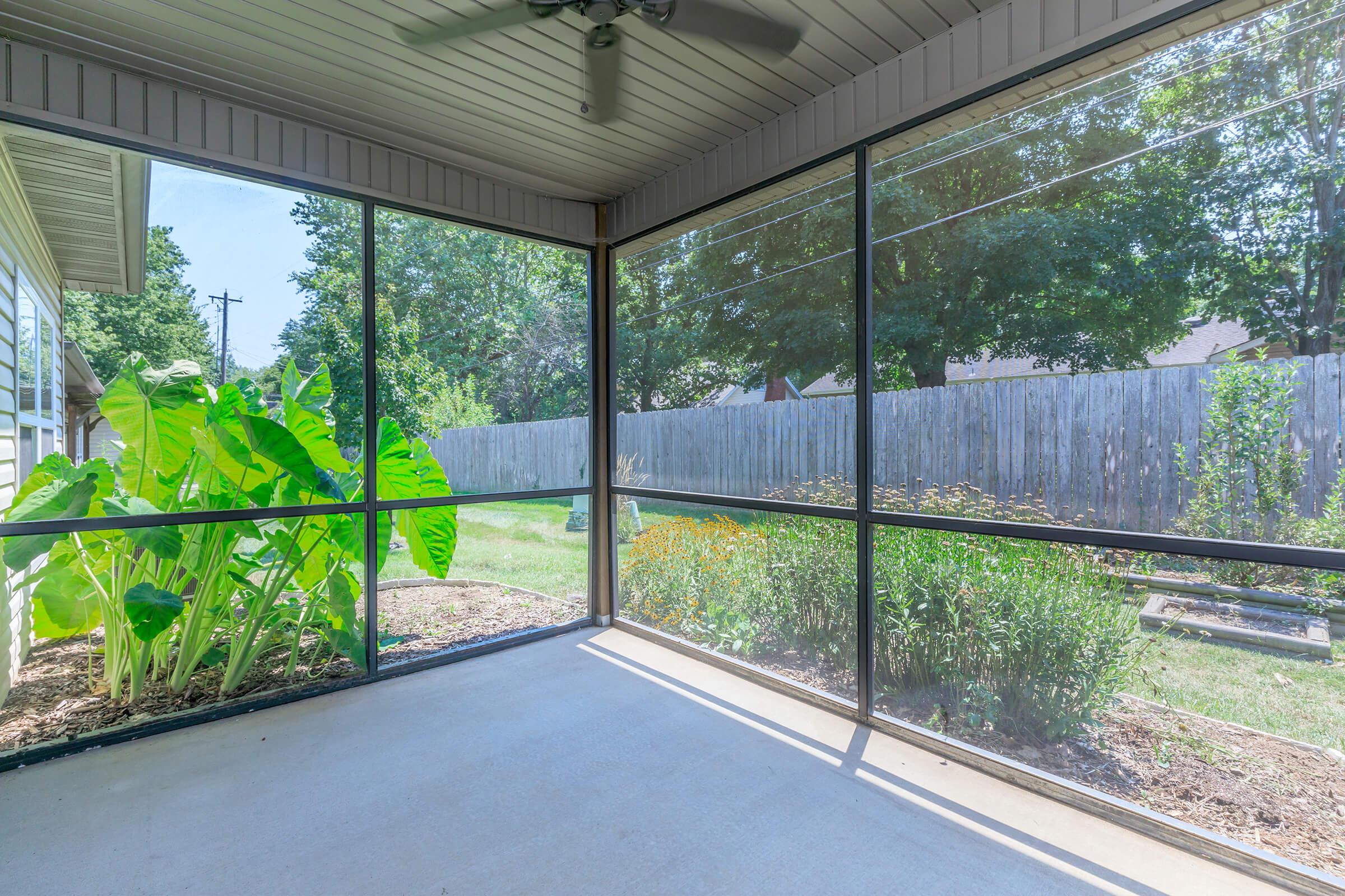 A screened porch with a concrete floor, featuring large green plants and a view of a backyard with trees and a wooden fence. Natural light filters in, creating a bright and open space for relaxation.