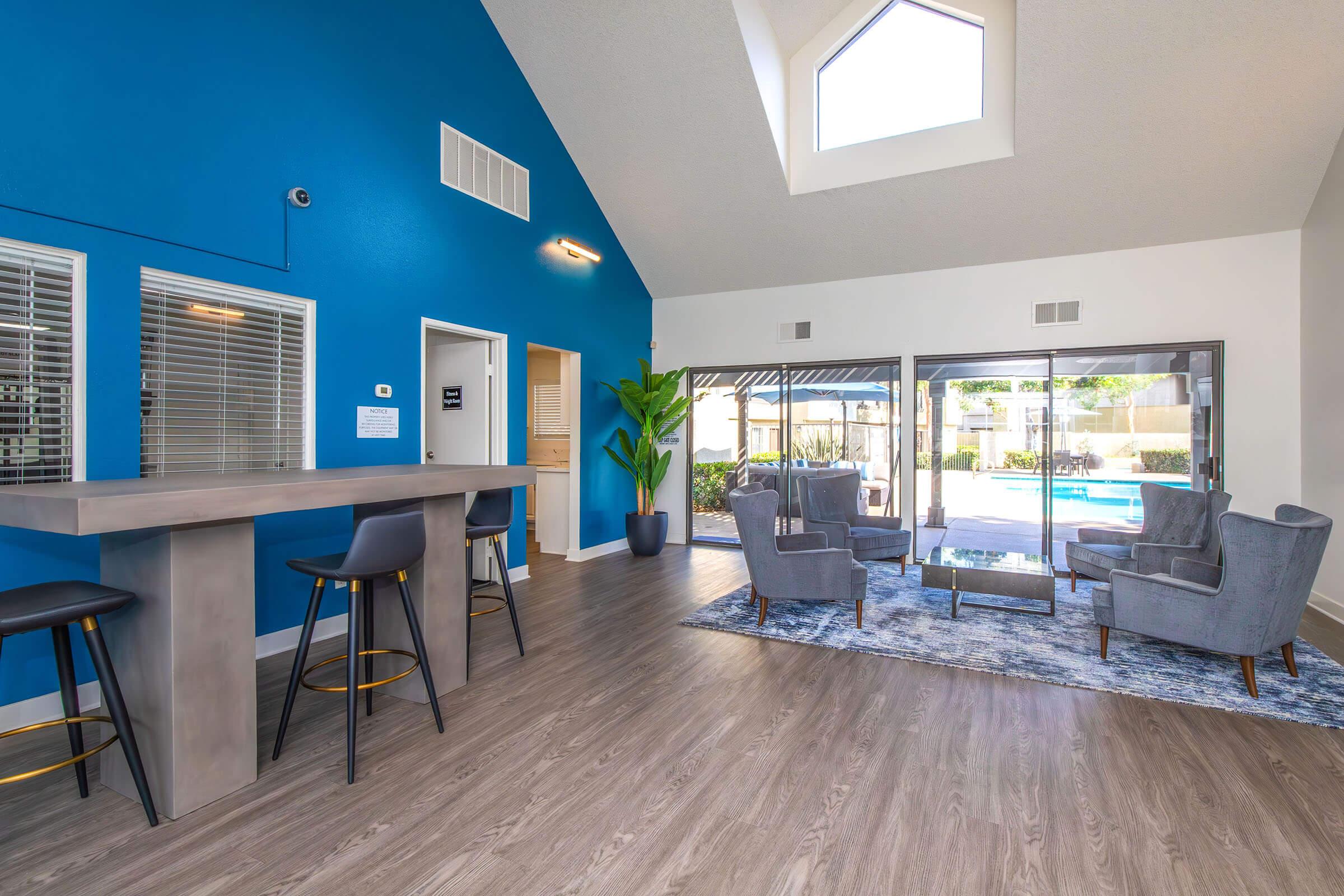 Interior of a modern community lounge featuring a blue accent wall, tall ceilings with a skylight, and large glass doors that open to an outdoor pool area. Furnished with gray chairs and a stylish rug, the space includes a bar height table with stools, creating a welcoming atmosphere.