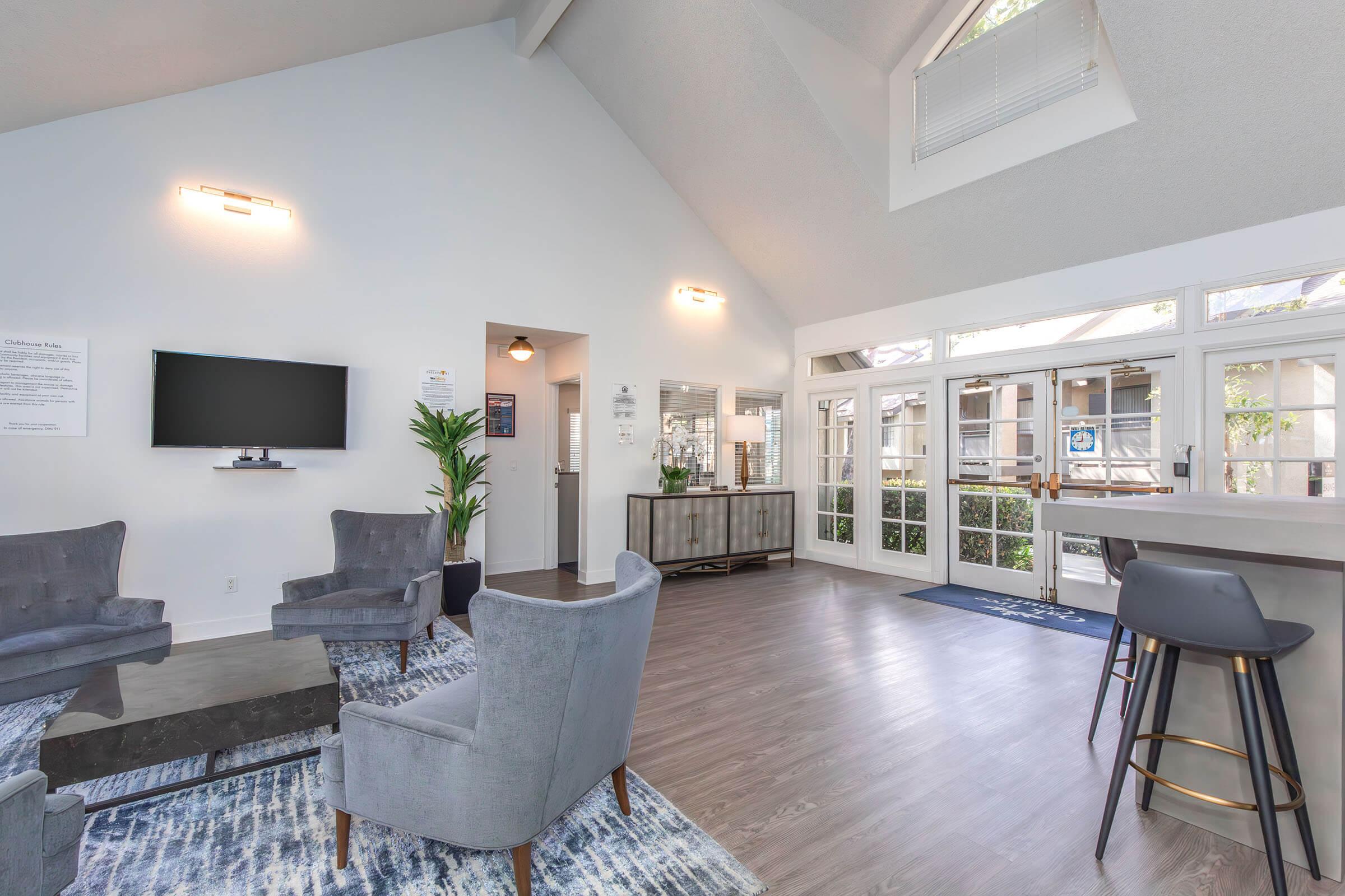 A modern, well-lit living area featuring two gray armchairs and a dark coffee table on a patterned rug. A wall-mounted TV is visible, along with a console table and a plant. Large windows provide natural light, and the space has an open, welcoming atmosphere.