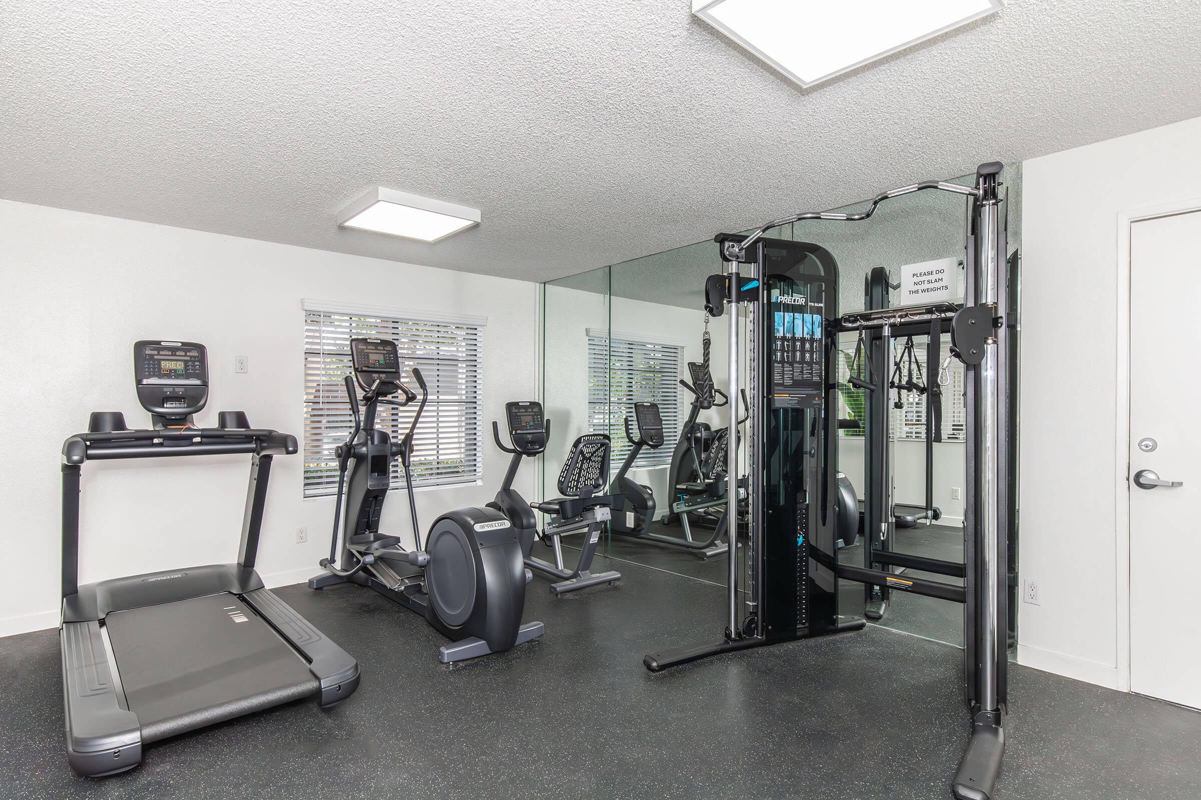 A fitness room featuring various exercise equipment, including a treadmill, elliptical machine, and multi-gym station. The space is well-lit with overhead lighting and has mirrors on one wall, creating a spacious feel. Windows allow natural light to enter, enhancing the workout environment.