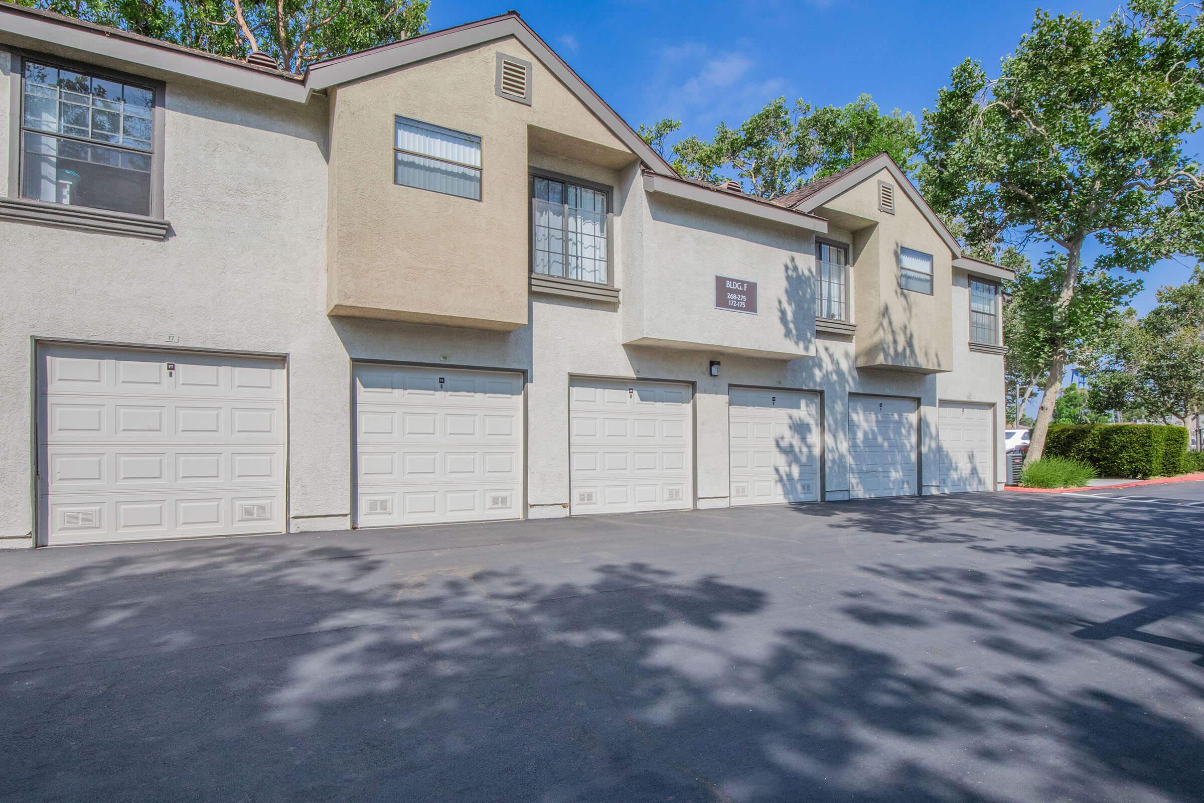 Row of tan and cream residential garages with closed doors, set against a clear blue sky. Shadows of trees fall on the pavement, creating a dappled light effect. The buildings feature large windows and simple architectural details, contributing to a suburban neighborhood appearance.