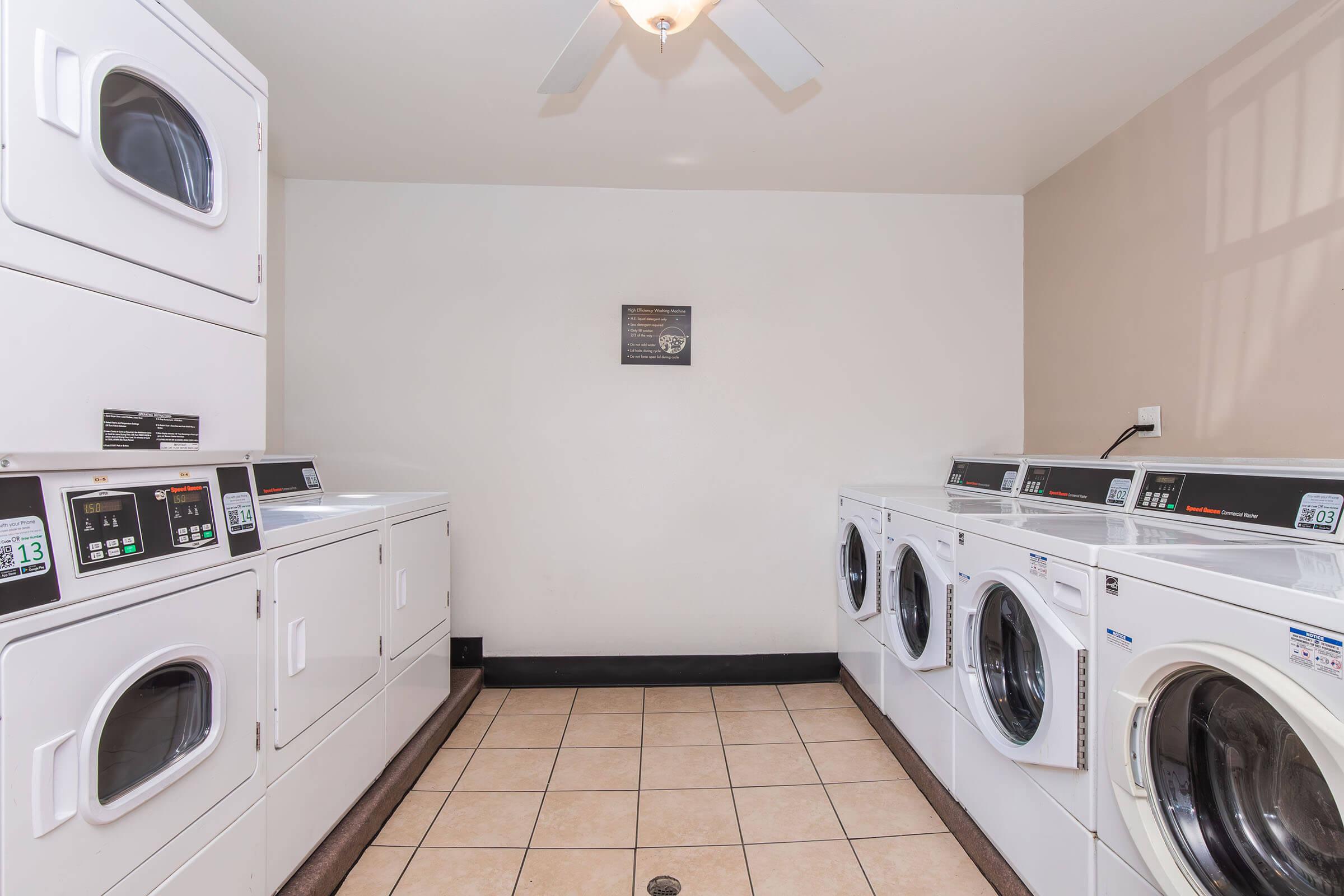 A clean laundry room featuring several white washers and dryers lined up against the walls. The floor is tiled, and there is a ceiling fan overhead. A small framed sign is displayed on the wall, adding a decorative touch to the functional space.