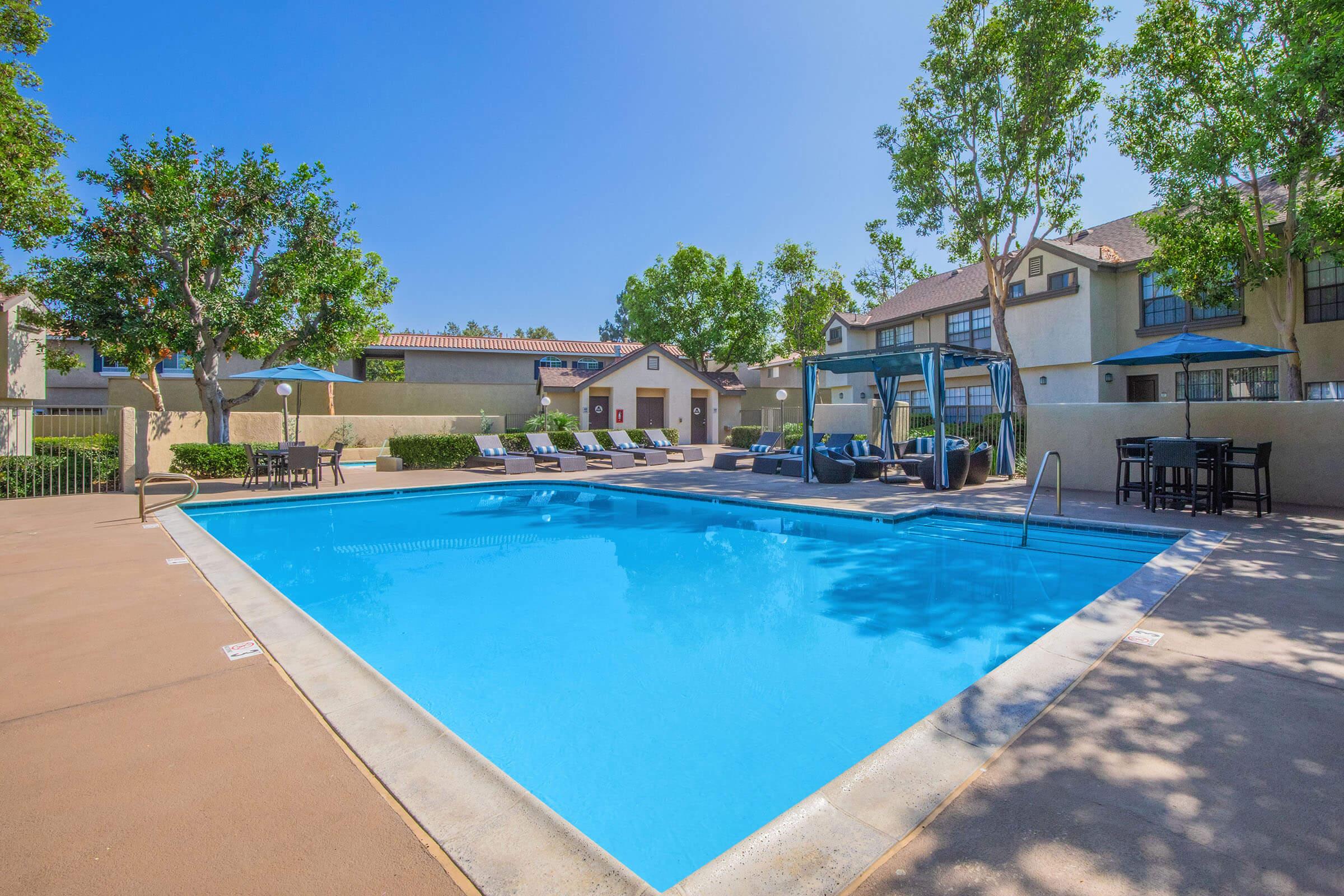 A bright and inviting swimming pool surrounded by lounge chairs and shaded seating areas. Trees and landscaping create a relaxing atmosphere, with residential buildings in the background. The clear blue water reflects the sunny sky, making it an ideal spot for leisure and relaxation.