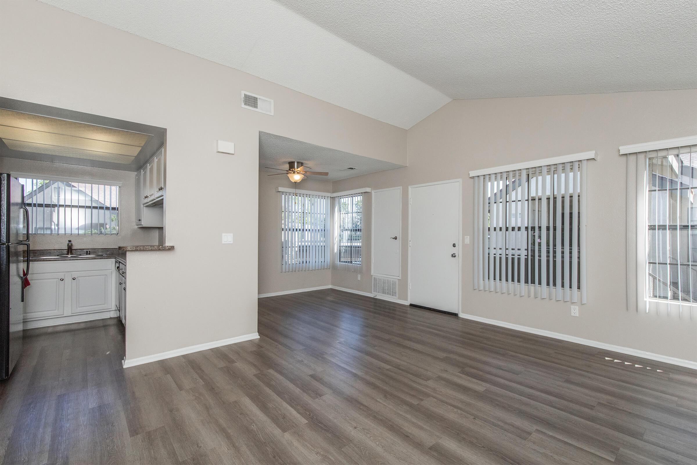 Bright and airy living space featuring an open floor plan. The room includes a kitchen area with white cabinets, a ceiling fan, and large windows with vertical blinds. Light-colored walls and laminate flooring enhance the spacious feel. A door leads to an outdoor area, providing natural light.