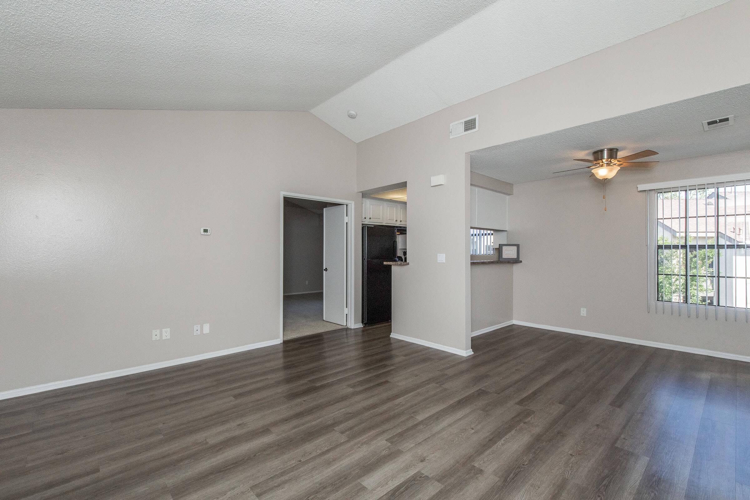 Spacious living area with light-colored walls, vaulted ceiling, and wood-like flooring. A door leads to a connected kitchen area, visible through an open doorway. Large windows allow natural light to illuminate the space, enhancing the inviting atmosphere.