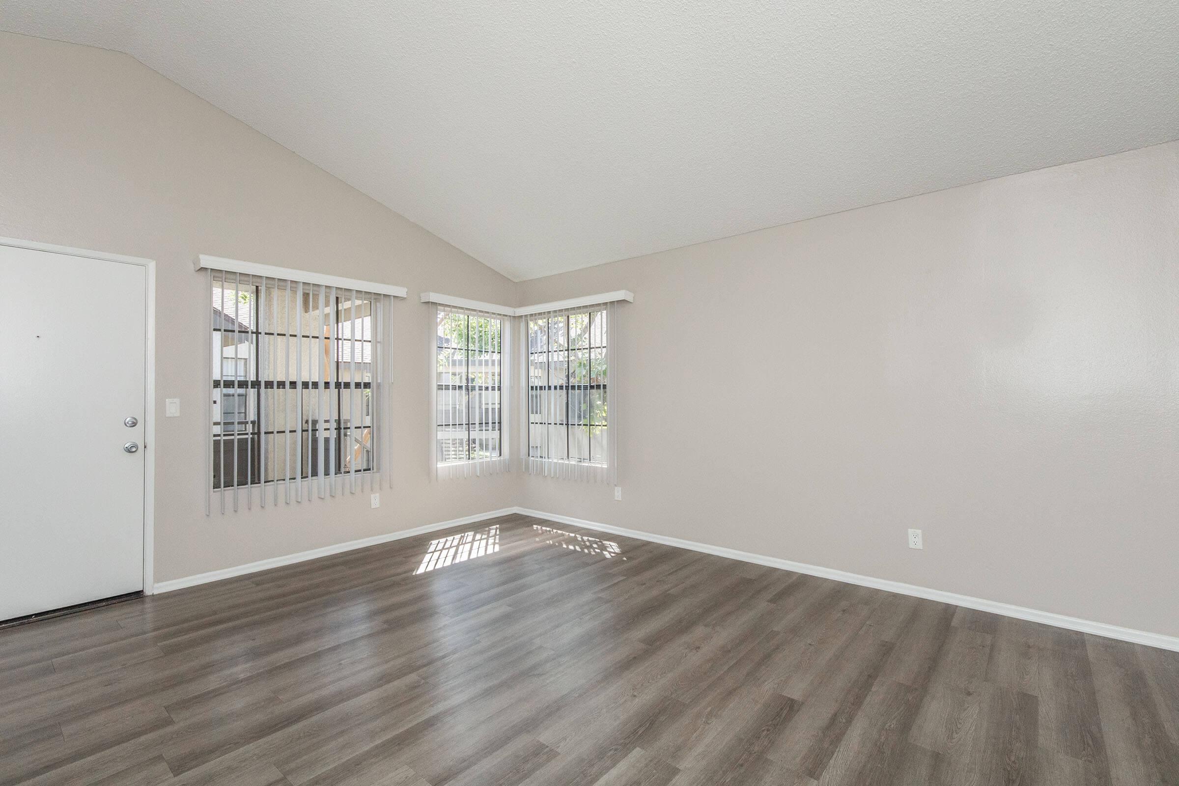 Spacious interior of a living room with high ceilings and large windows allowing natural light. The walls are painted in a light color, and there is a modern laminate floor. Vertical blinds cover the windows, and the entry door is located on the left side of the image.