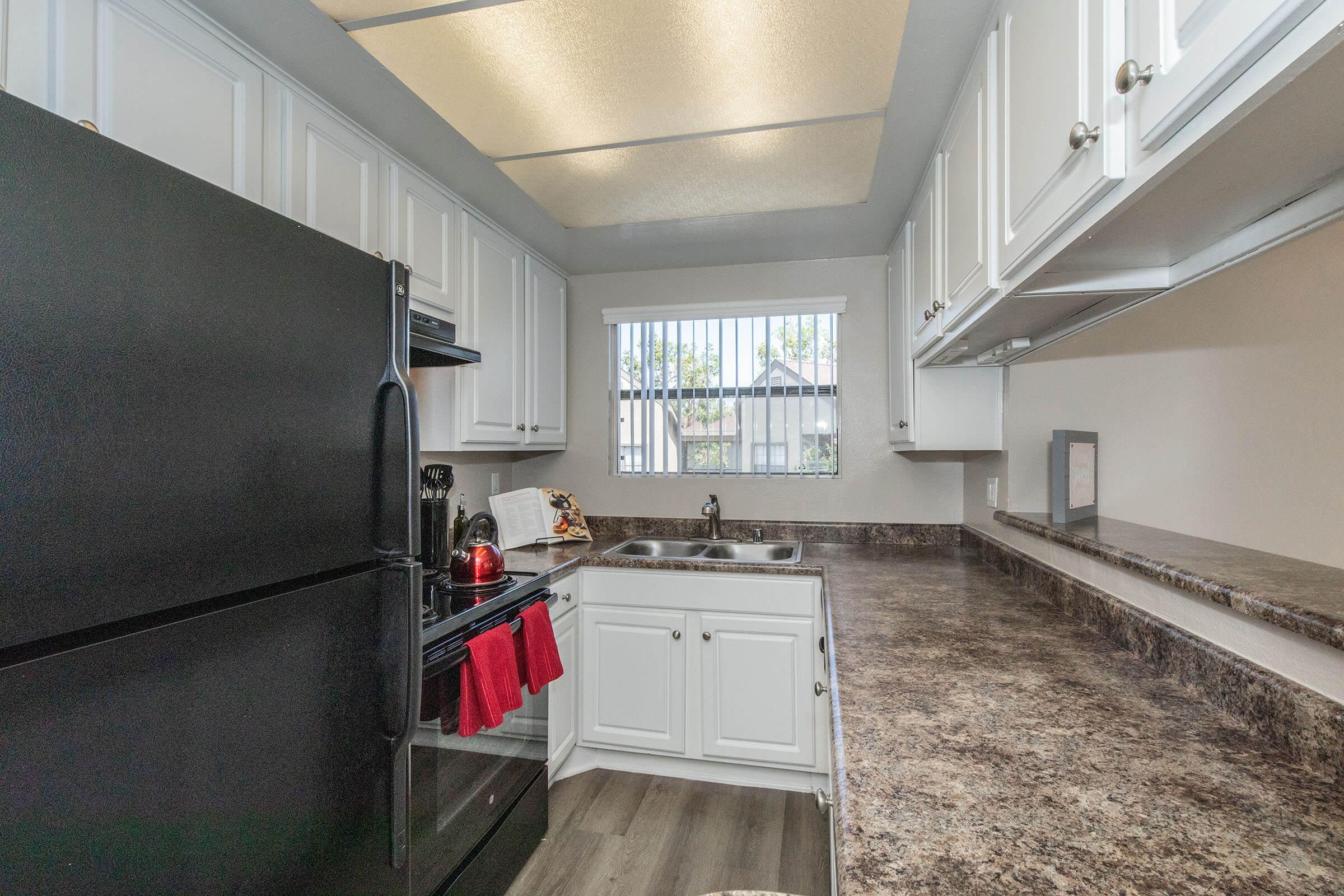 A modern kitchen featuring a black refrigerator, white cabinets, and a dark countertop. A sink is integrated into the counter, with a window providing natural light. The space includes kitchen appliances, a red dish towel hanging, and a few decorative items on the counter.