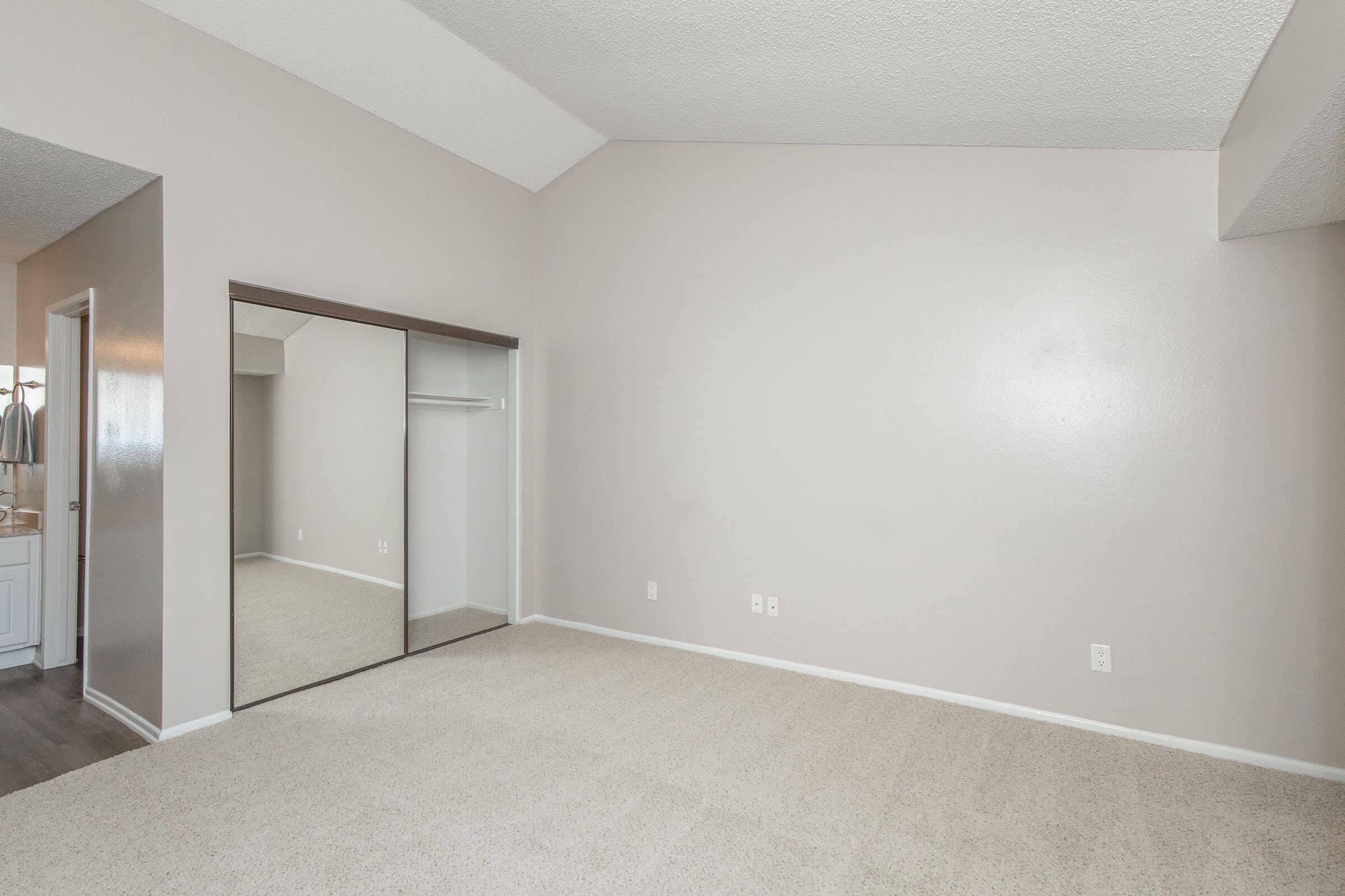 A spacious, empty room with light-colored walls and carpeted floor. It features a large mirror closet on one side and an adjoining doorway leading to another room. The ceiling is vaulted, creating a sense of openness. Natural light may enter through unseen windows.