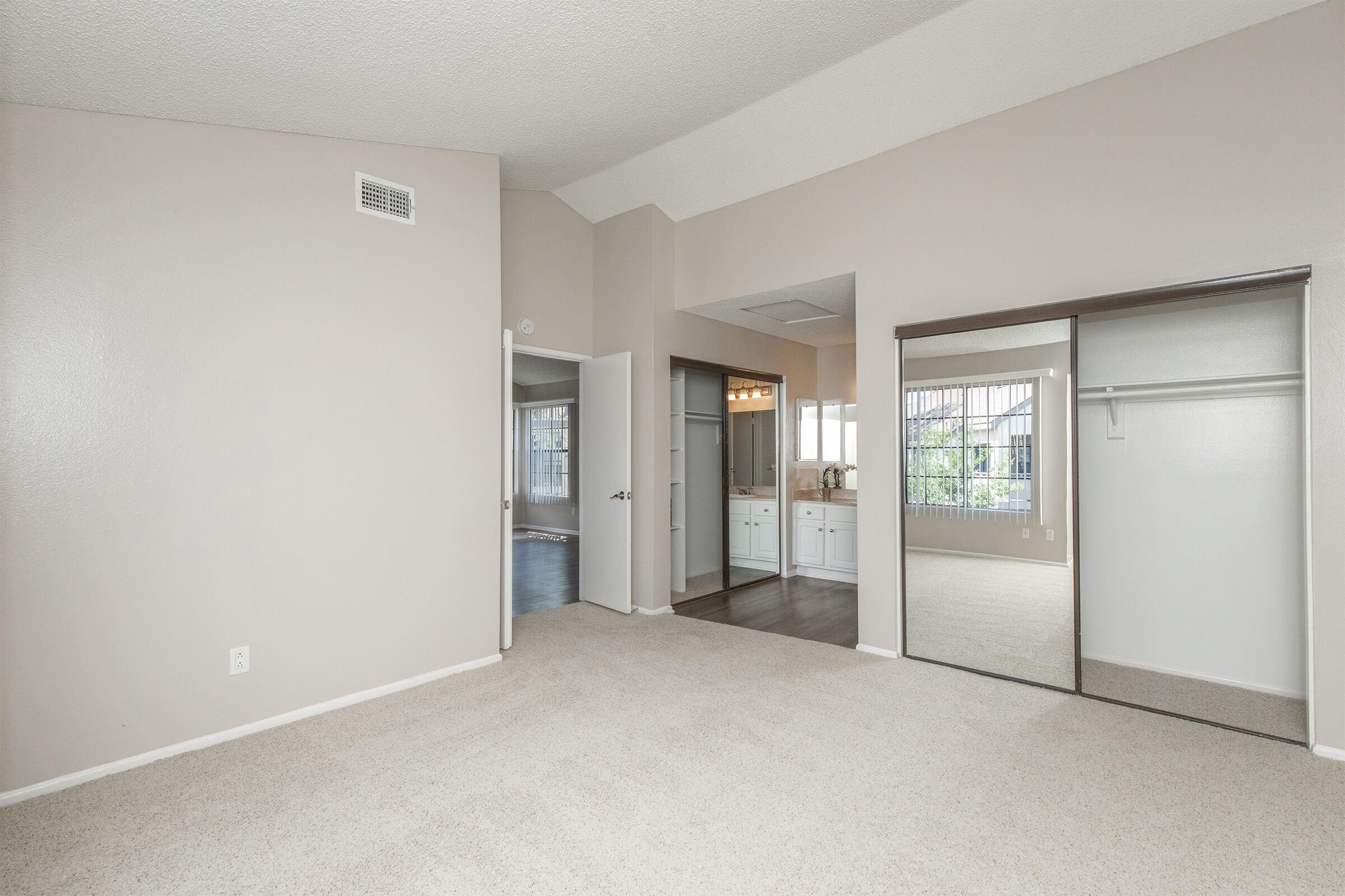 A spacious room featuring light-colored walls and carpet. There are mirrored closet doors on one side, and a door leading to a bathroom area in the background. Bright natural light enters through windows, enhancing the inviting atmosphere. The ceiling is high, creating an open feel.