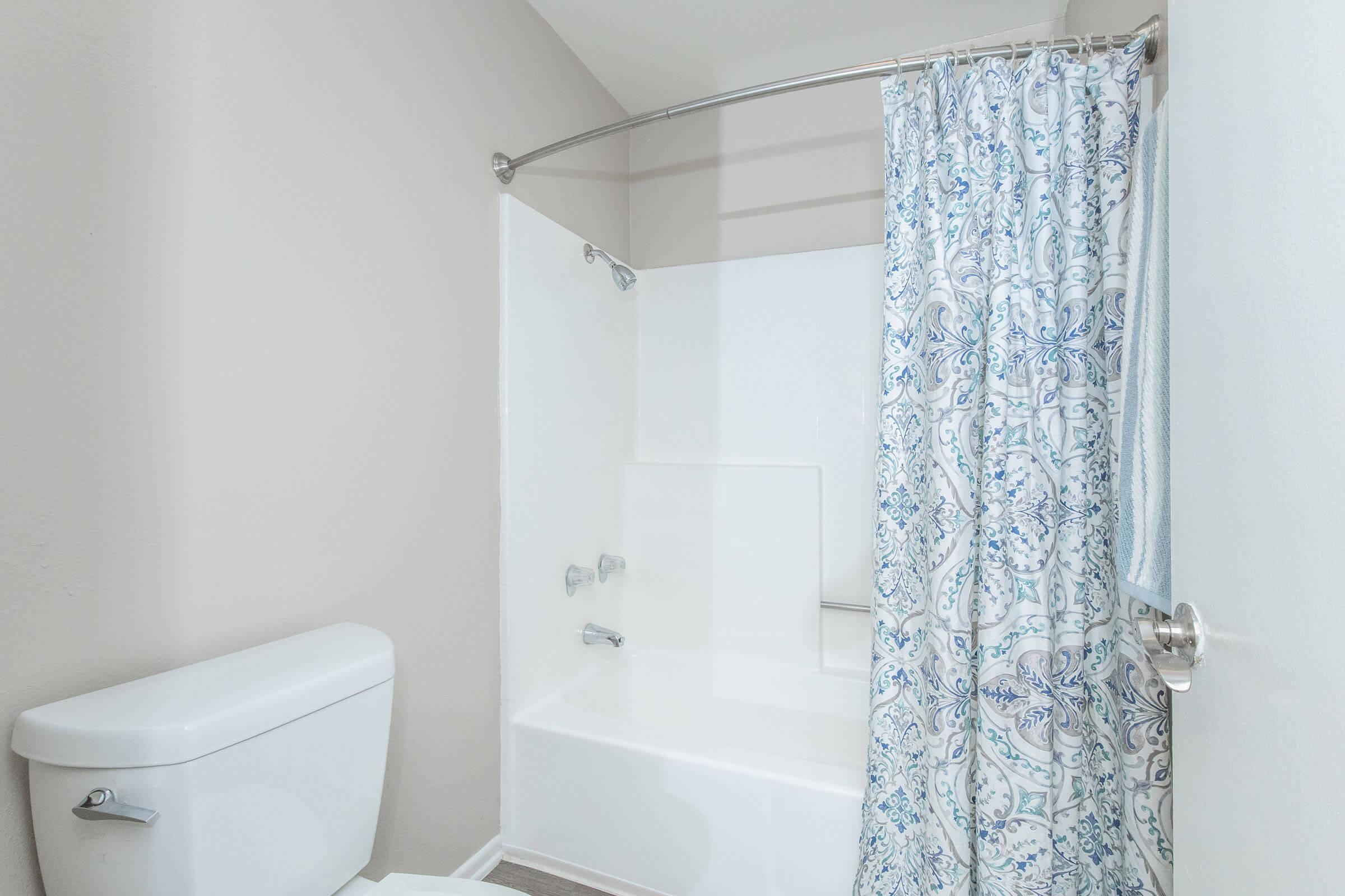 A clean and modern bathroom featuring a bathtub with a shower curtain displaying a blue and white floral pattern. There's a toilet adjacent to the bathtub, and the walls are painted in a light, neutral color. The overall design is simple and inviting.
