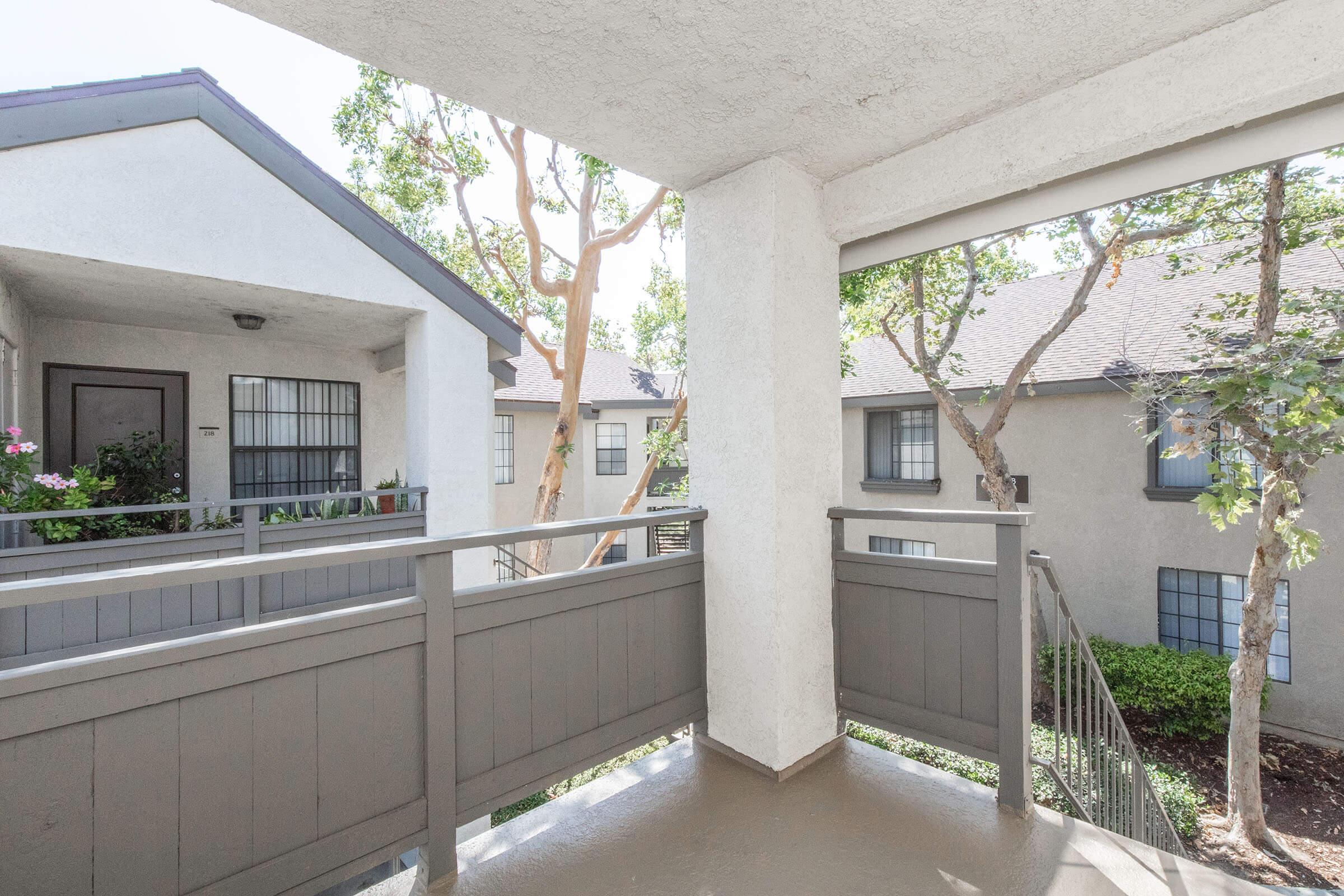 View of a balcony with gray railings overlooking a landscaped area. The scene includes trees and nearby buildings with windows. Bright daylight enhances the atmosphere, creating a serene outdoor space. Potted plants are visible on the balcony, adding a touch of greenery.