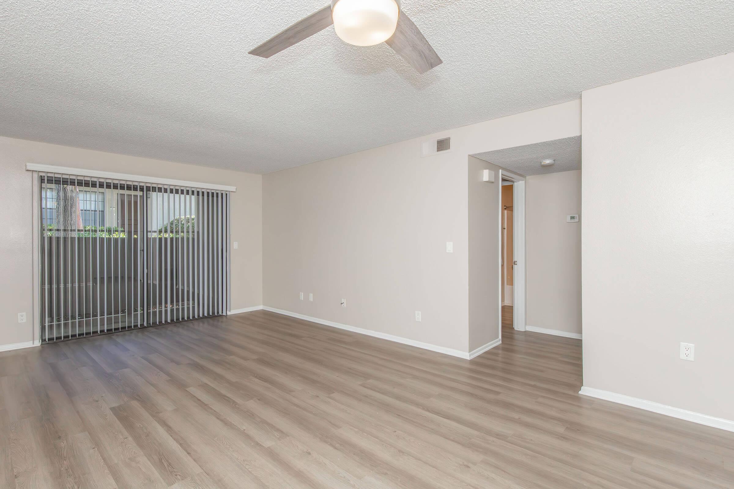 Spacious living area with light-colored walls and wood-like flooring, featuring a ceiling fan. Large sliding glass doors on the left, leading to a patio, let in natural light. An archway on the right leads to another room, creating an open and airy atmosphere.