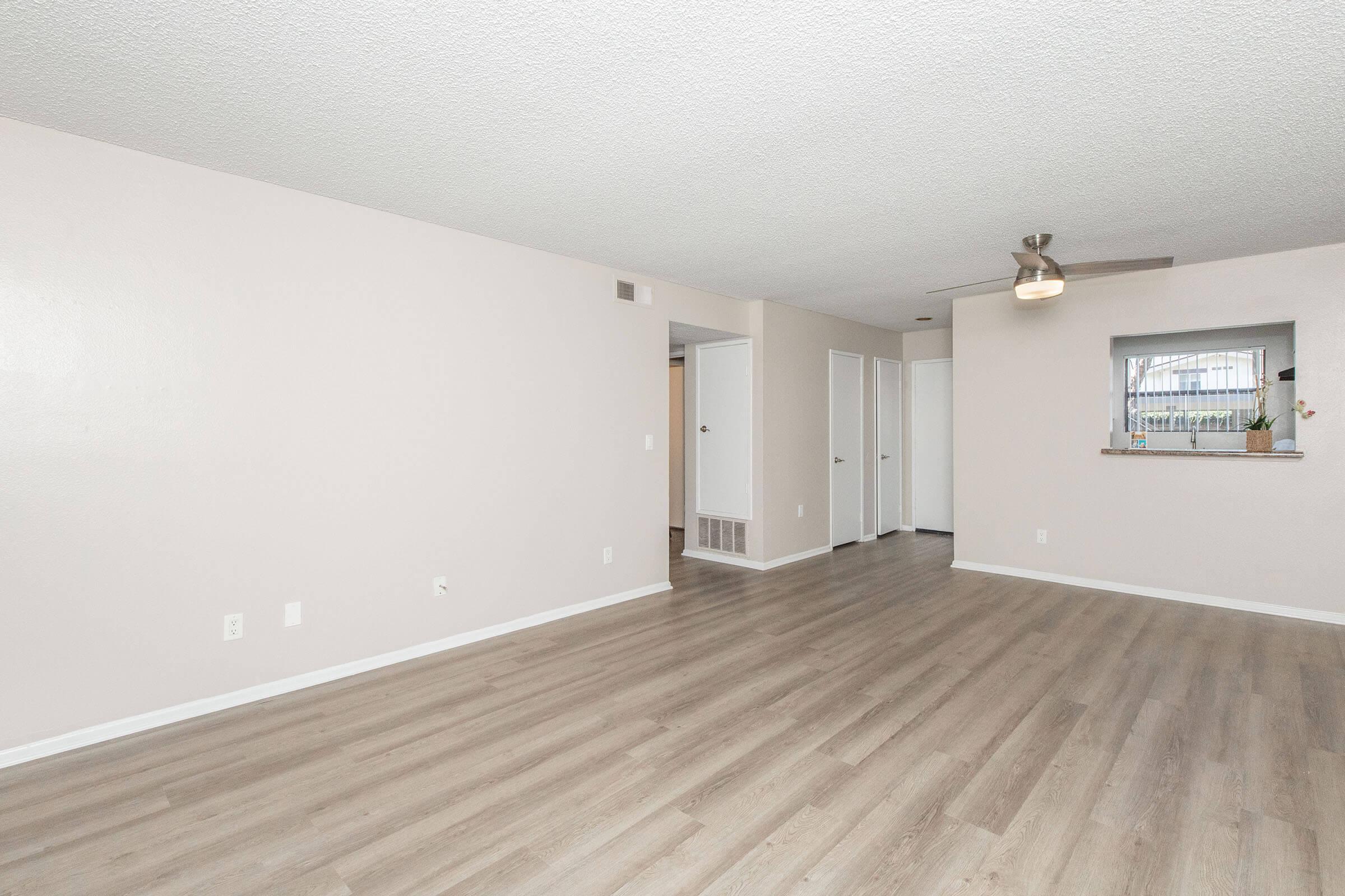A spacious, unfurnished room with light-colored walls and wooden flooring. There is a ceiling fan and a cut-out window leading to another area. Multiple doorways are visible, leading to adjacent rooms, creating a bright and open atmosphere.