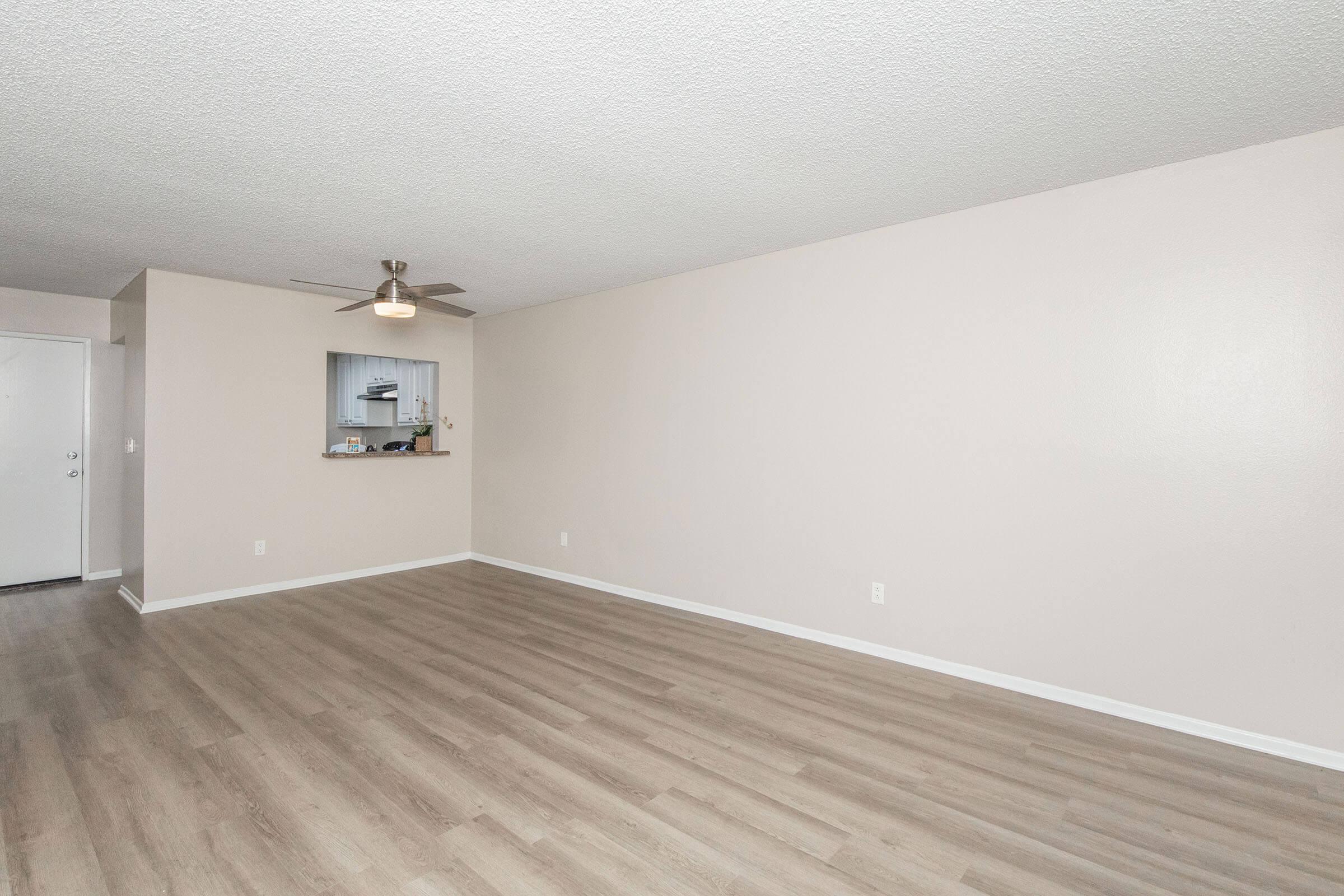 Empty living room featuring a light-colored wall, wooden floor, and a ceiling fan. An opening leads to a kitchen area in the background, with minimal decor and no furniture. Natural light illuminates the space, creating a bright and airy atmosphere.