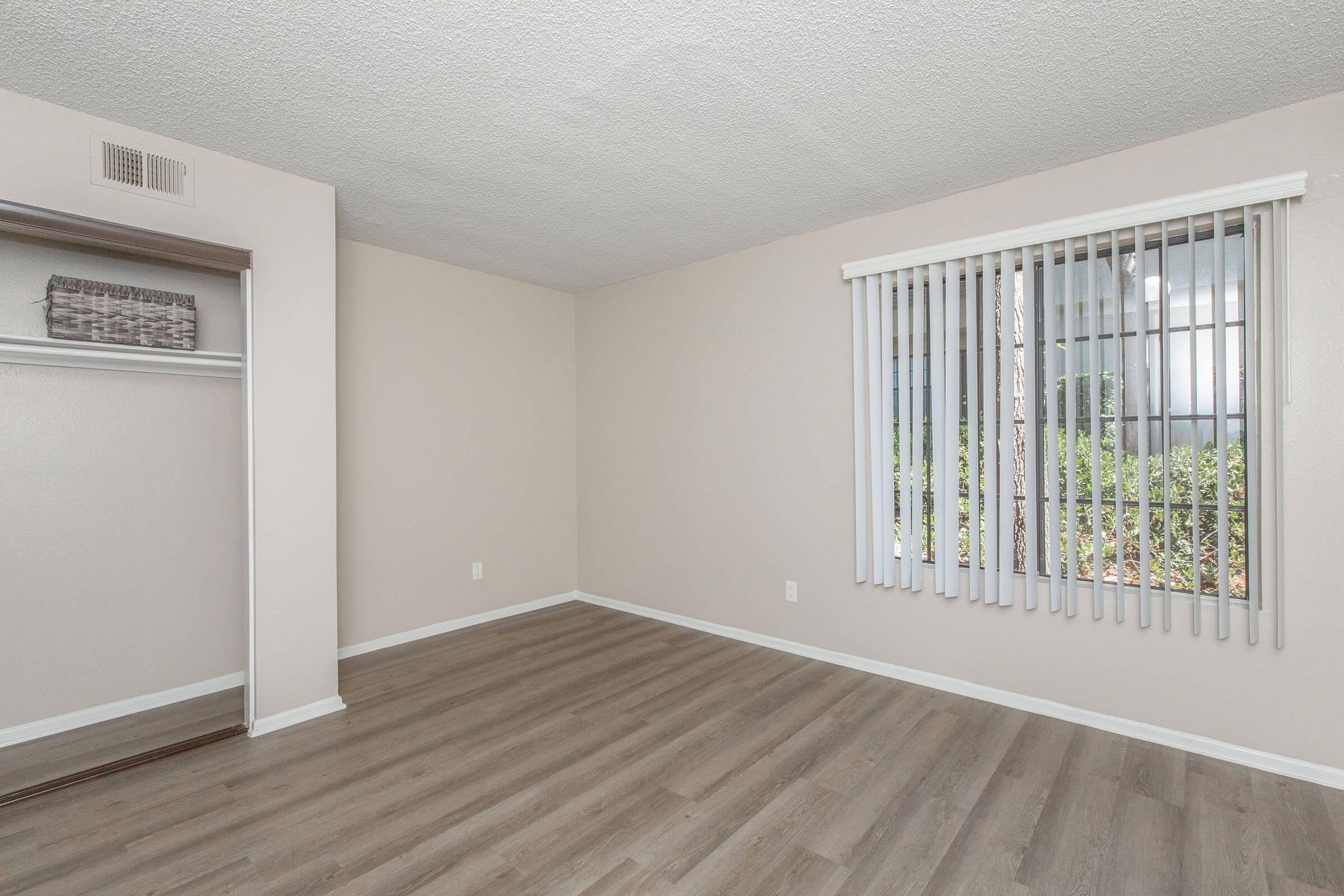 A spacious, empty room with light-colored walls and wooden laminate flooring. It features a window with vertical blinds, allowing natural light to enter, and a closet with sliding doors on one side. The room is clean and ready for furnishing, providing a blank canvas for decoration.
