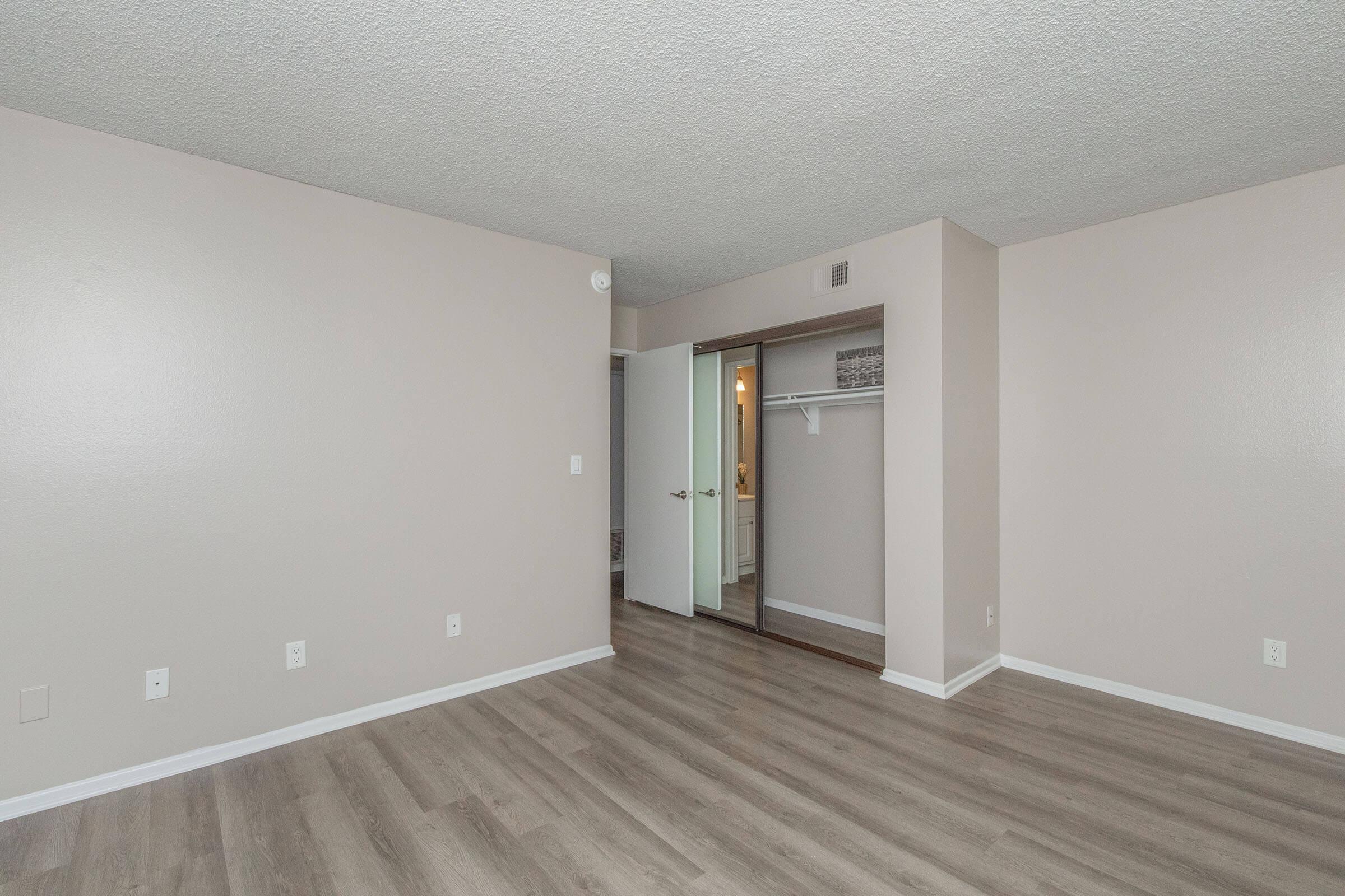 Empty living space featuring light-colored walls and new flooring. A closet with sliding mirrored doors is visible to the right, and an open door leads to another area. The room is well-lit with a neutral color palette, creating a spacious and inviting atmosphere. Ideal for new occupants to personalize.