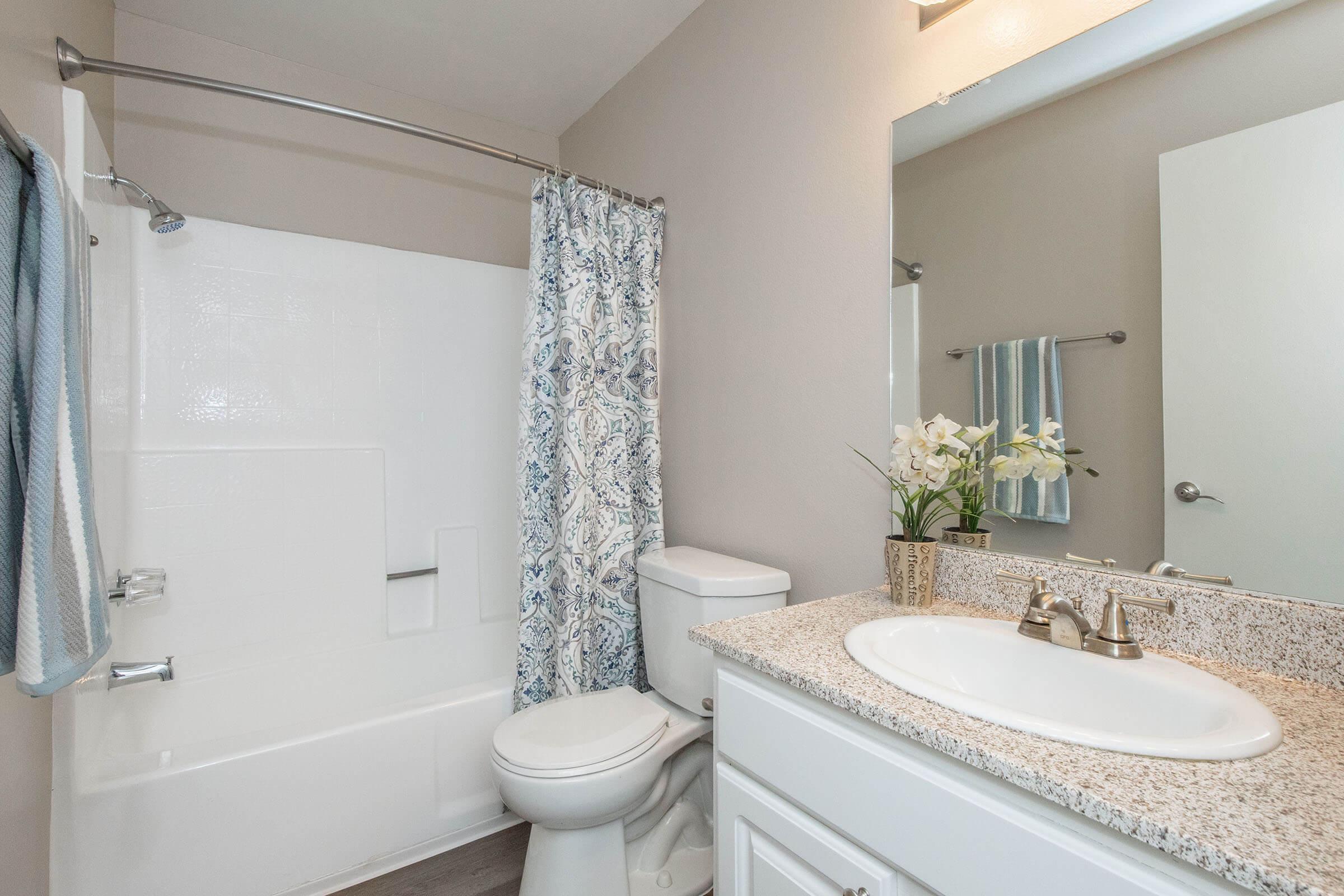A clean and modern bathroom featuring a bathtub with a shower curtain, a white toilet, and a vanity with a sink. The countertop has a decorative vase of white flowers, and there's a blue towel hanging on a rack. The walls are painted in a neutral tone, creating a bright and inviting space.