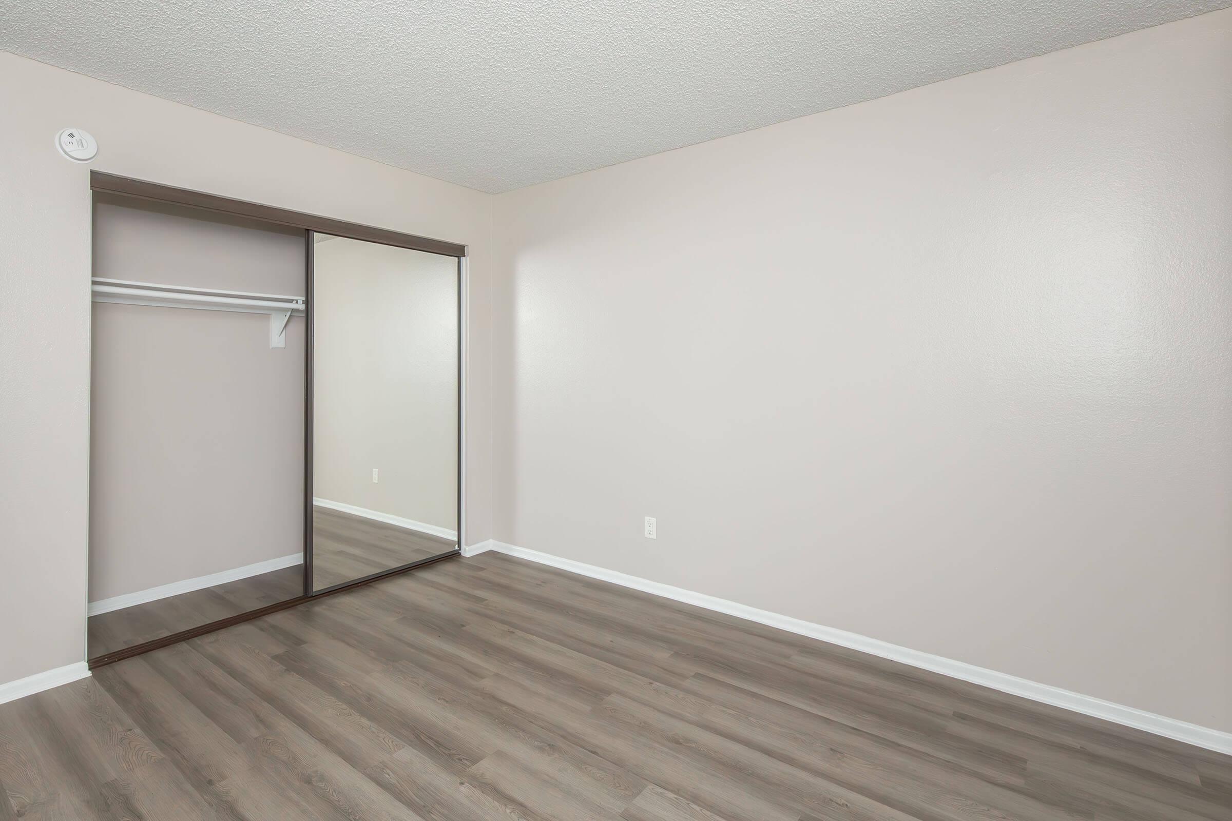 A lightly colored, empty bedroom with a smooth wooden floor. There's a mirrored closet with sliding doors on the left side, and the walls are painted a soft beige. The room is well-lit and spacious, offering a blank canvas for decoration.
