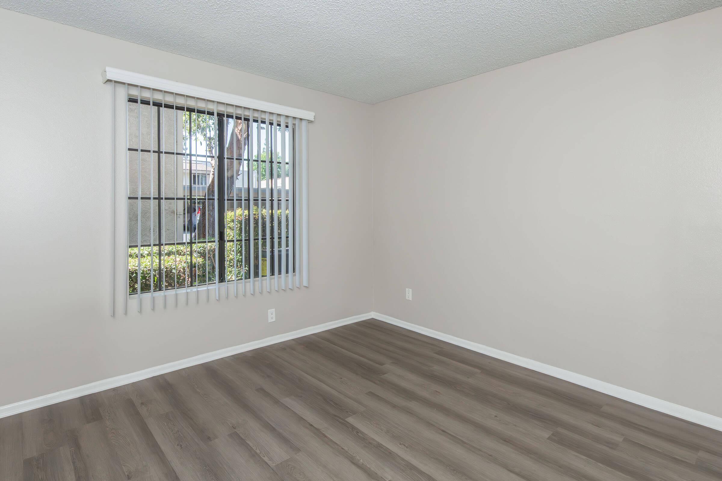 Empty room with light beige walls and wooden flooring. A large window with vertical blinds lets in natural light, showing greenery outside. No furniture or decorations are present, creating a clean and spacious feel.