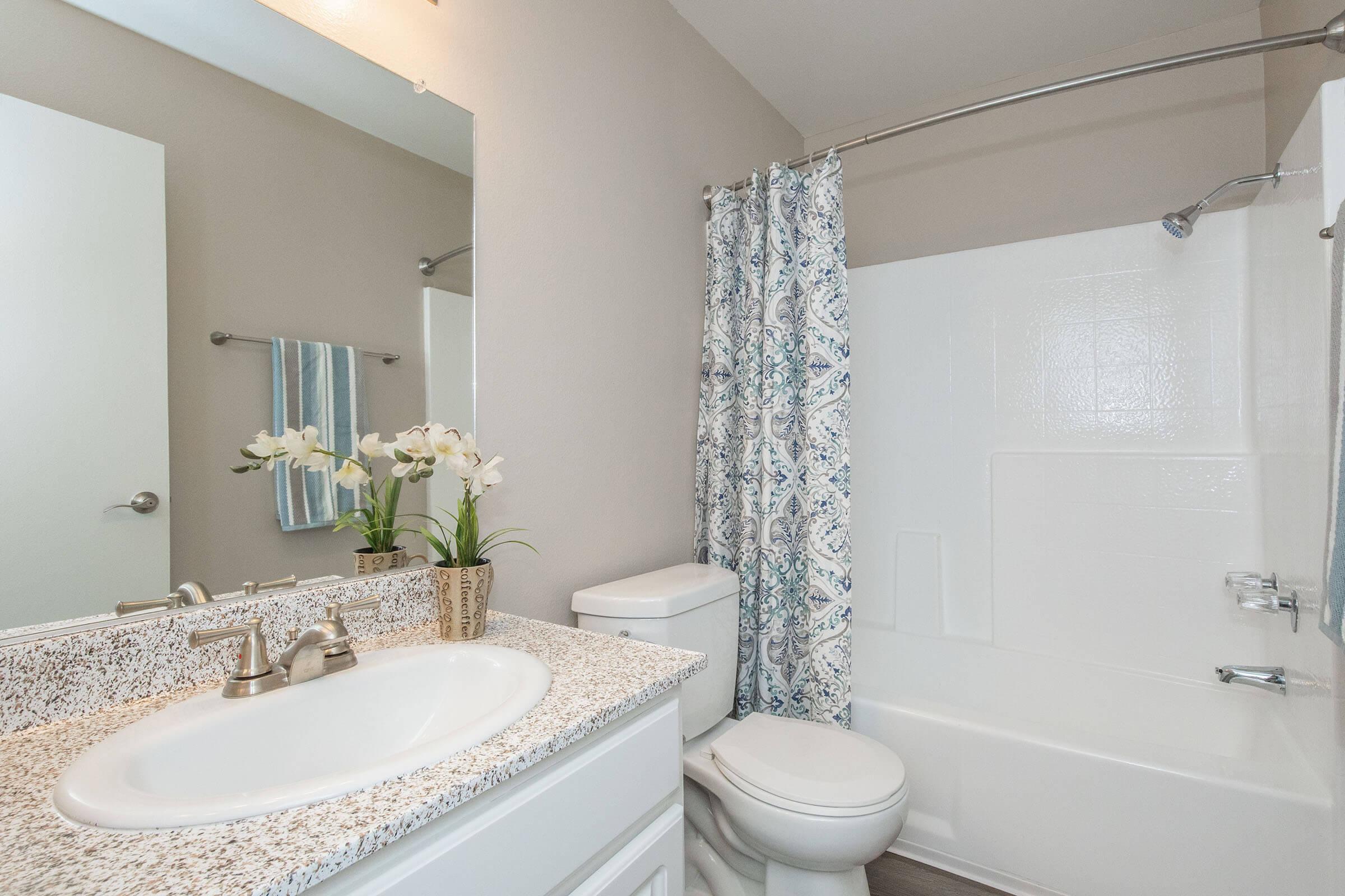 A modern bathroom featuring a white bathtub and shower, a toilet, and a sink with a countertop. The walls are painted in a light color, and a decorative curtain with a blue and white pattern hangs by the shower. A small vase with white flowers sits on the countertop, adding a touch of elegance.