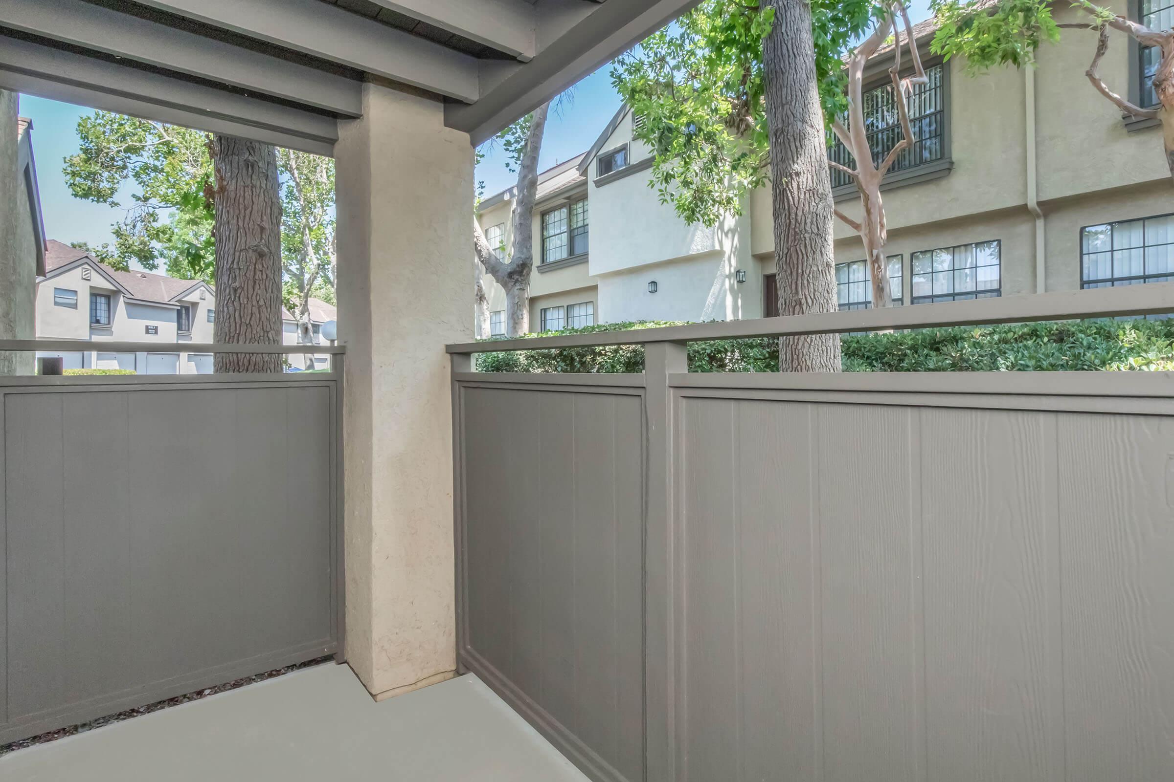 A view from a covered porch or balcony showing a gray wooden railing, with green trees and neighboring buildings visible in the background. The space is bright and inviting, framed by the architectural features of the porch.