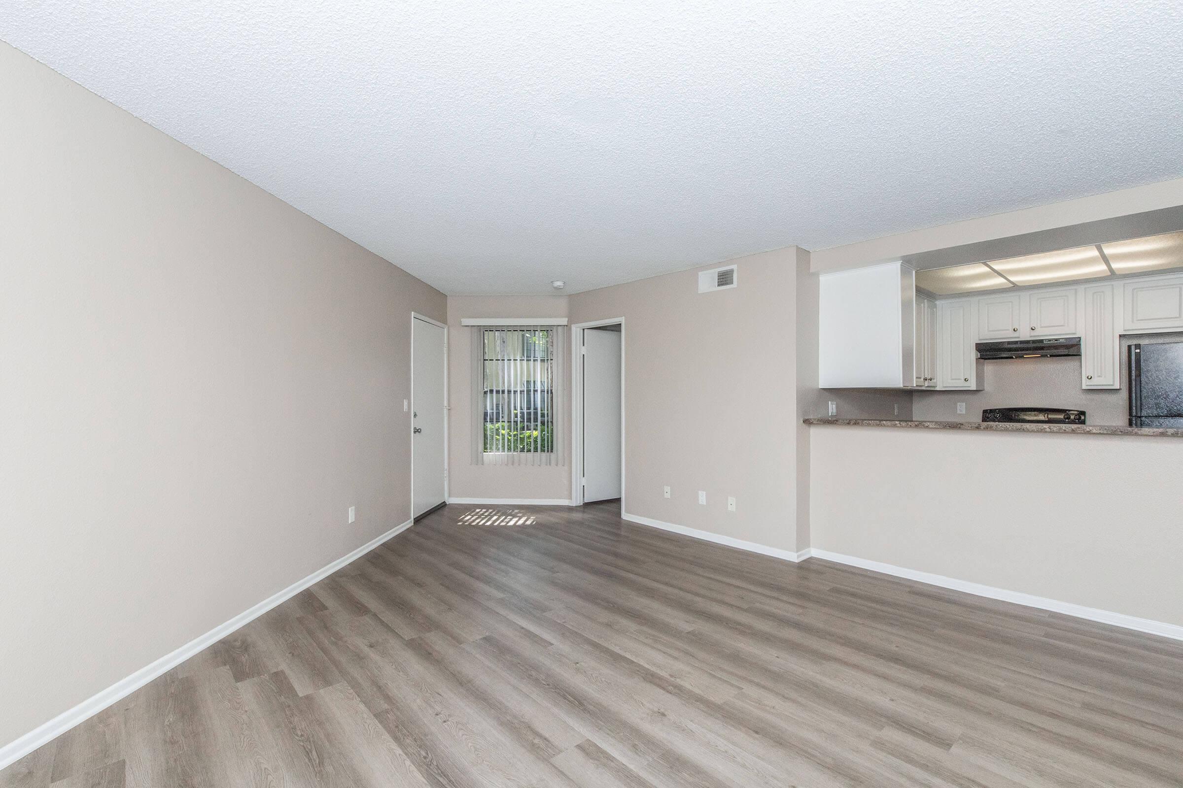 Spacious, well-lit living area with light-colored walls and modern wood-like flooring. Features an open layout connecting to a kitchen with white cabinetry. A large window allows natural light, and there are doors leading to an adjacent room. The space appears clean and inviting, ideal for comfortable living.