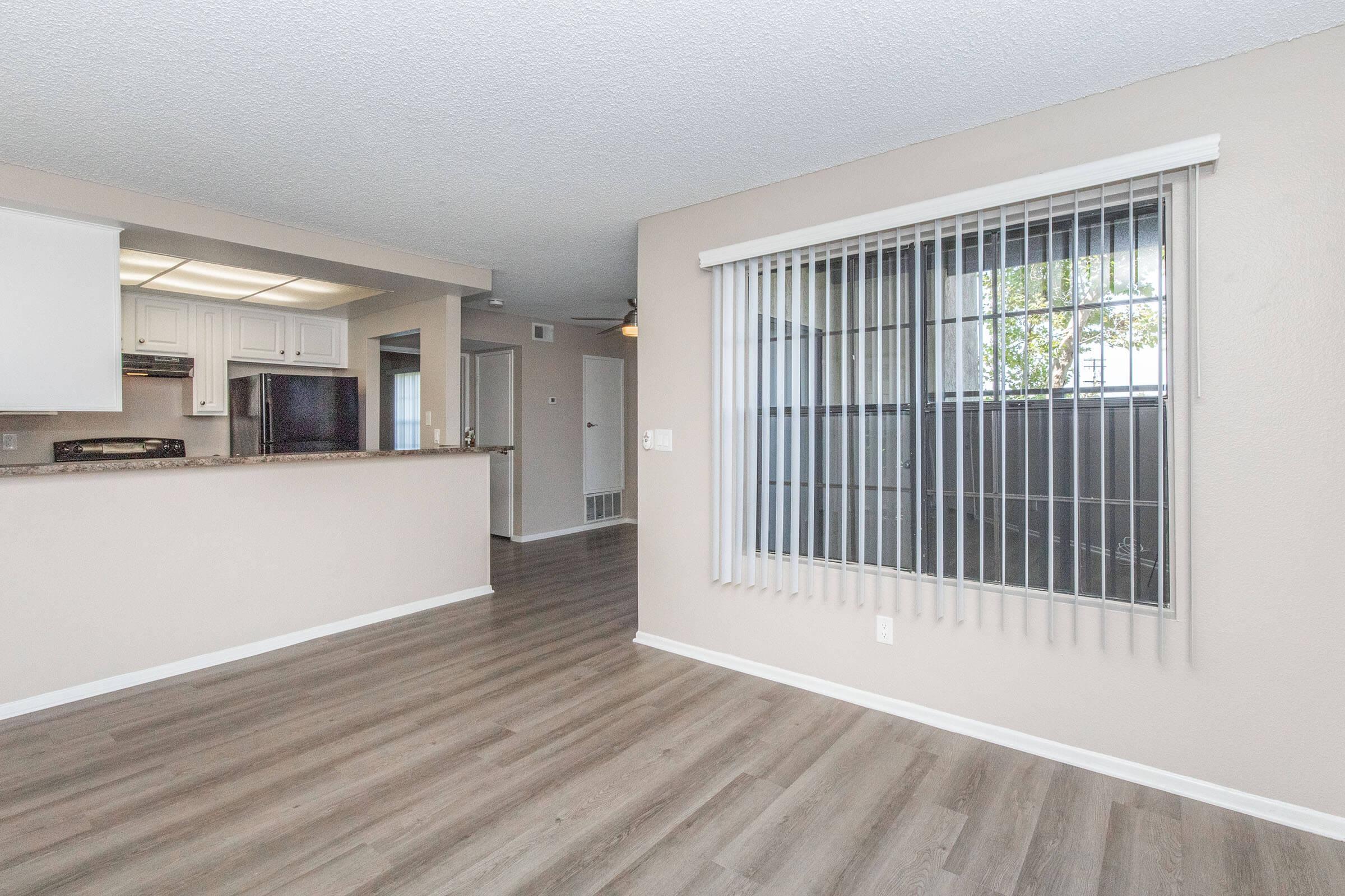 Interior view of a modern apartment featuring an open layout. The room has light-colored walls, large windows with vertical blinds, and laminate flooring. A kitchen is visible in the background with white cabinets and a granite countertop, creating a spacious and inviting atmosphere.
