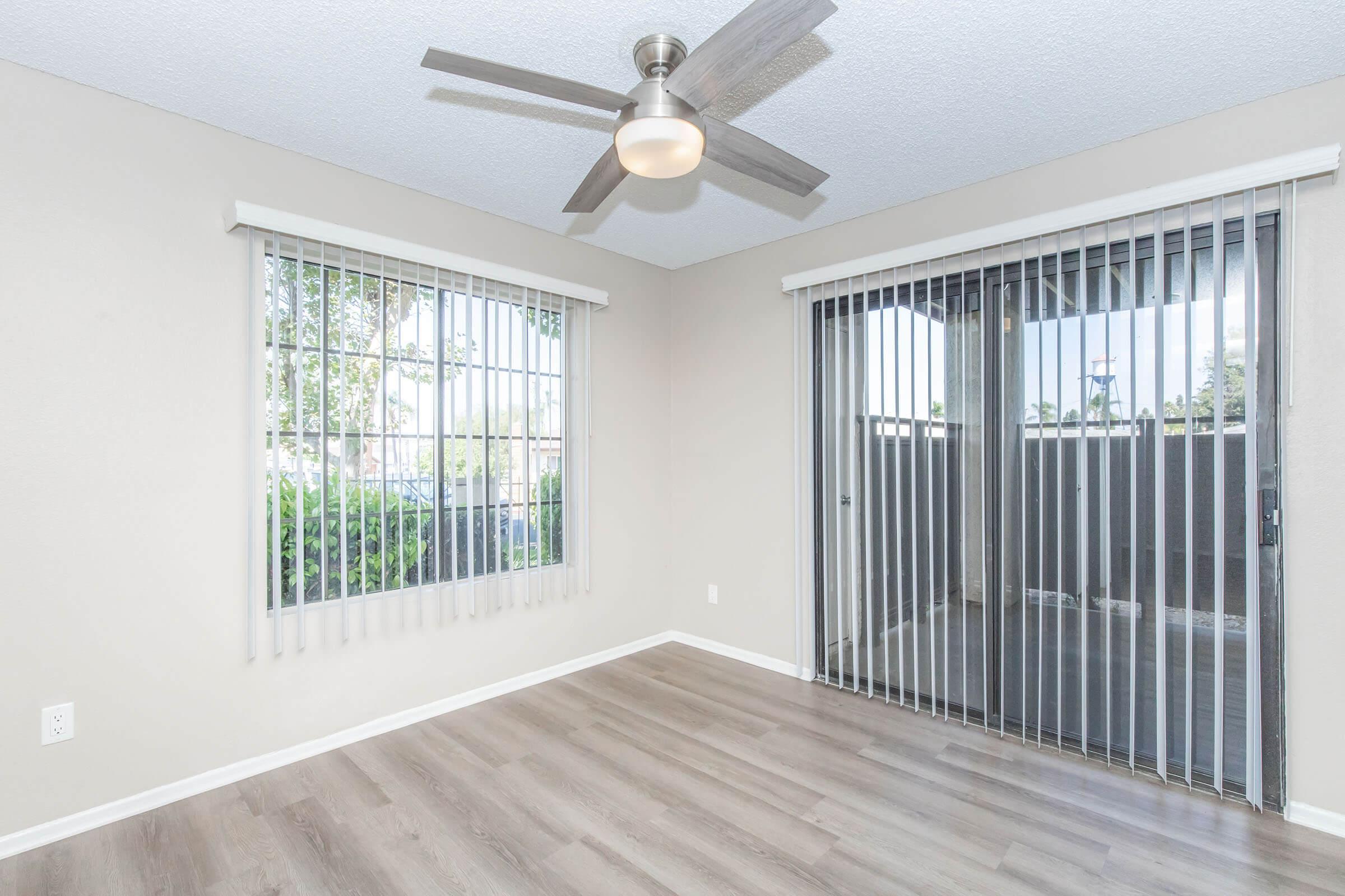 An empty room with light gray walls, wooden flooring, and a ceiling fan. It features two windows with vertical blinds, one overlooking a balcony. The space is well-lit and has a clean, modern appearance.