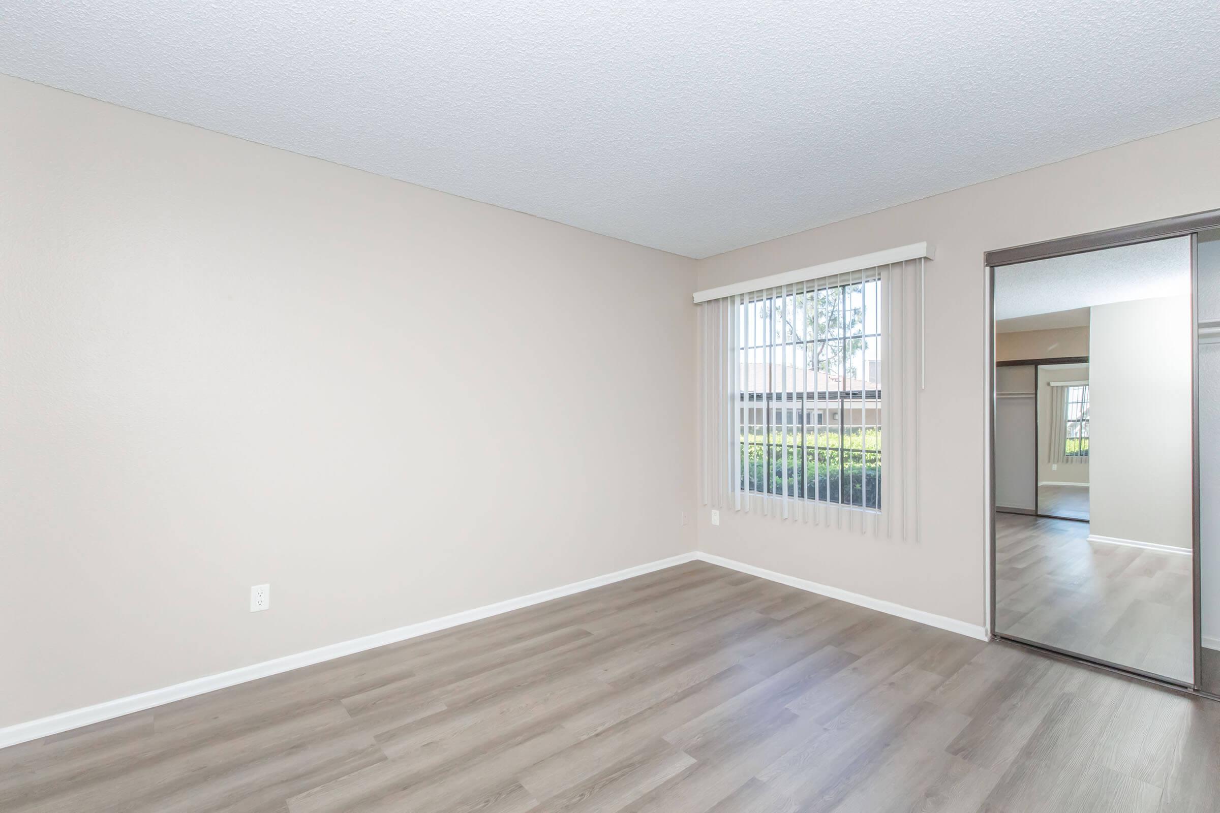 Empty room with light beige walls and vinyl flooring. A large window with blinds lets in natural light, and a mirrored closet door is on one side. The space feels open and airy, suitable for various furnishing options.