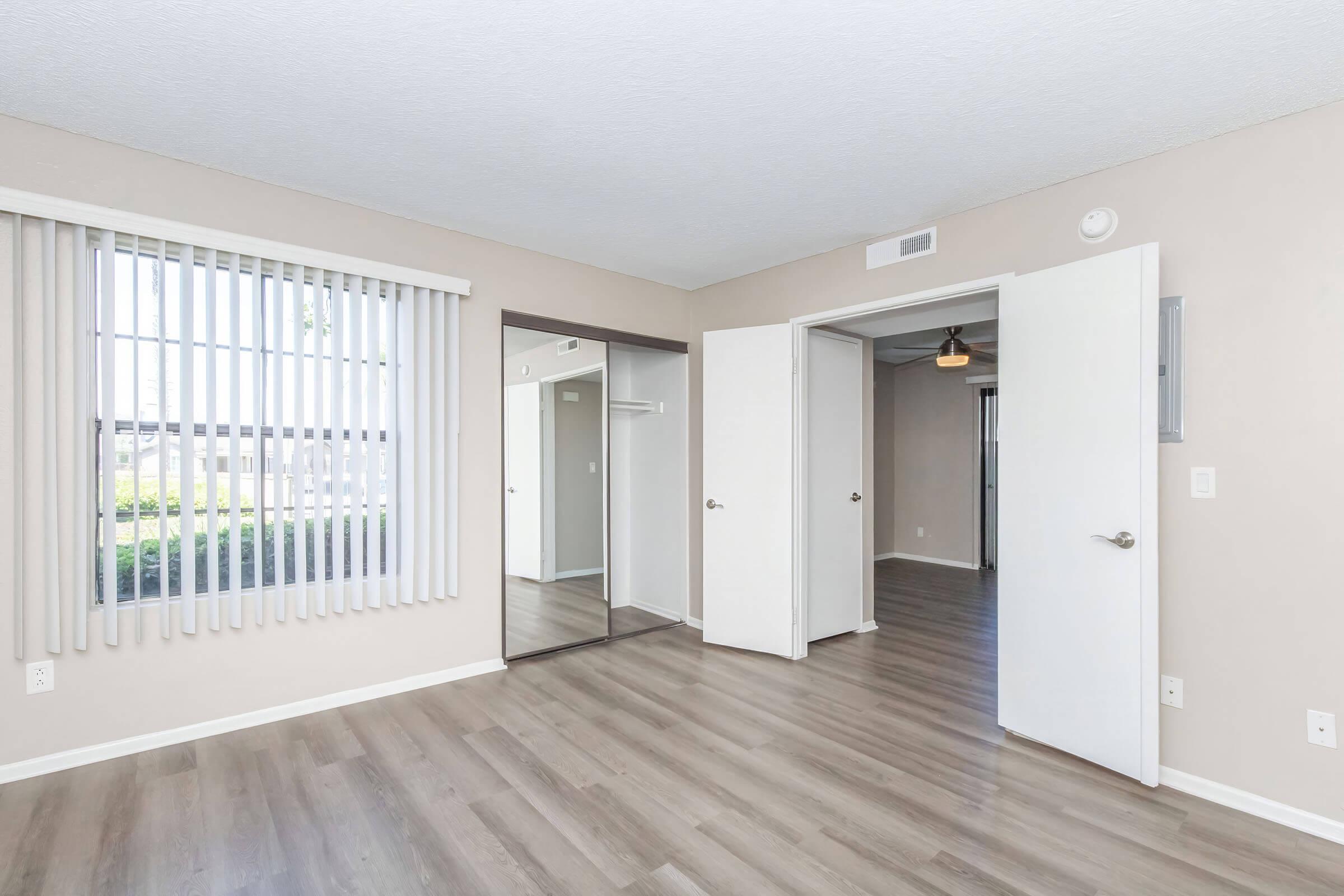 A spacious interior room featuring light beige walls, laminate flooring, and large windows with vertical blinds. The room includes two doors leading to different areas and a full-length mirror on one wall, enhancing the sense of space and natural light.