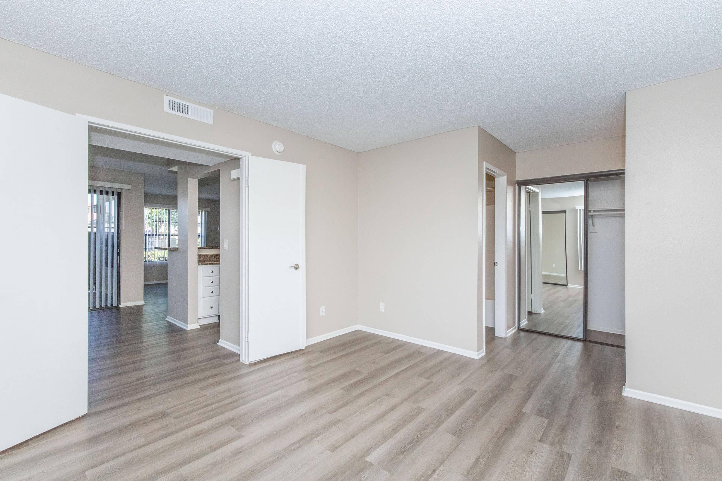 Interior view of a spacious room with light-colored walls and wooden flooring. Two doorways are visible, leading to other areas. There is a closet with sliding doors on one side. Natural light enters the space, creating a bright and open atmosphere.