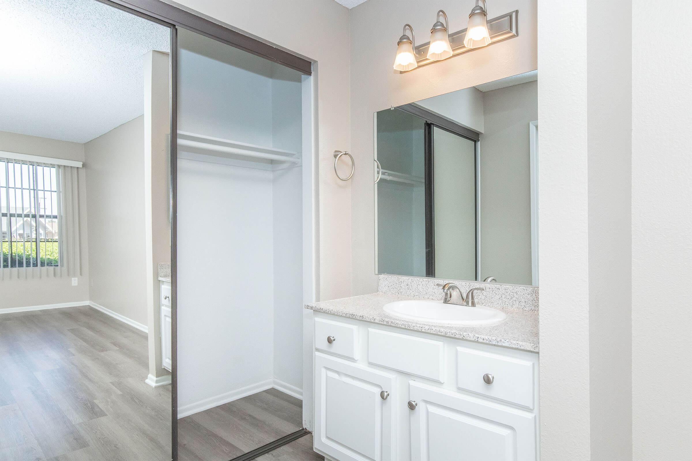A bathroom with a single vanity sink and mirror, featuring white cabinets and a light-colored countertop. An open closet with sliding doors is visible on the left. The wall is painted in a light color, and there's a window in the background, allowing natural light to enter the space. The flooring is a light wood pattern.