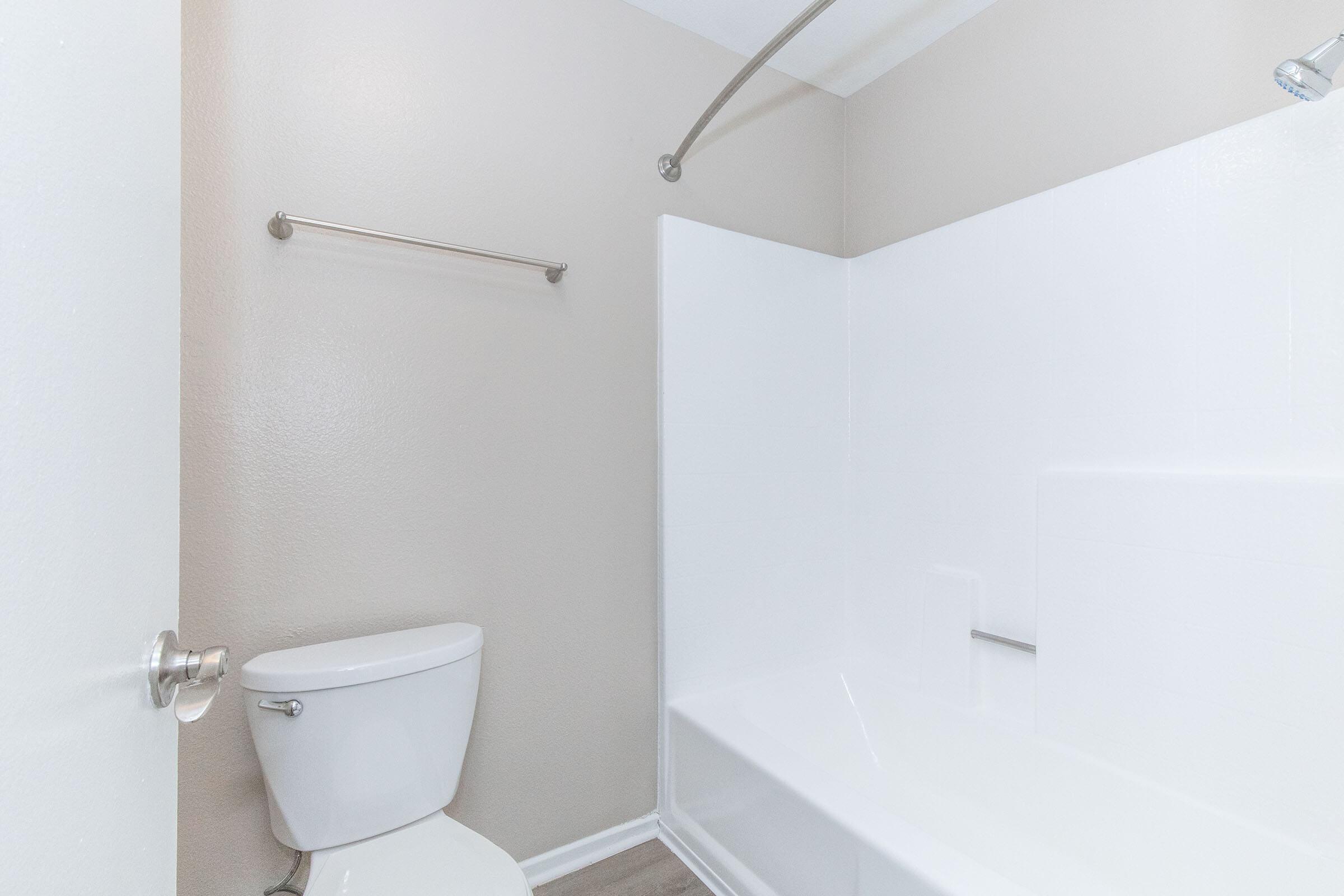 A clean, modern bathroom featuring a white bathtub with a curved shower rod, a toilet, and light beige walls. The floor displays a simple wood-like design, and the space is well-lit, creating a fresh and inviting atmosphere.