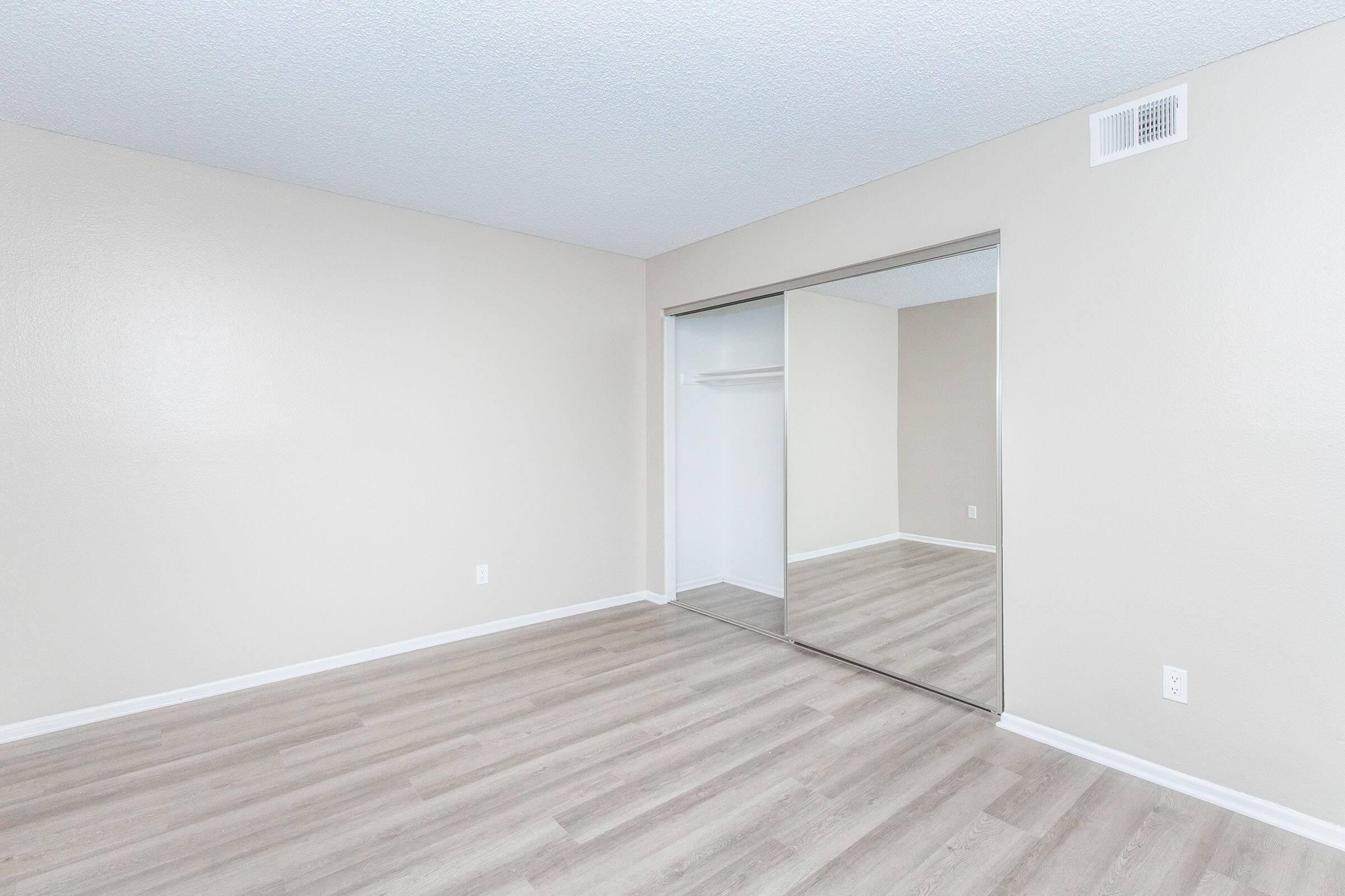 A vacant room with light beige walls and light wood-like laminate flooring. The room features a mirrored closet with sliding doors and a small white closet area. The space appears clean and modern, suitable for various uses, with no furniture present.