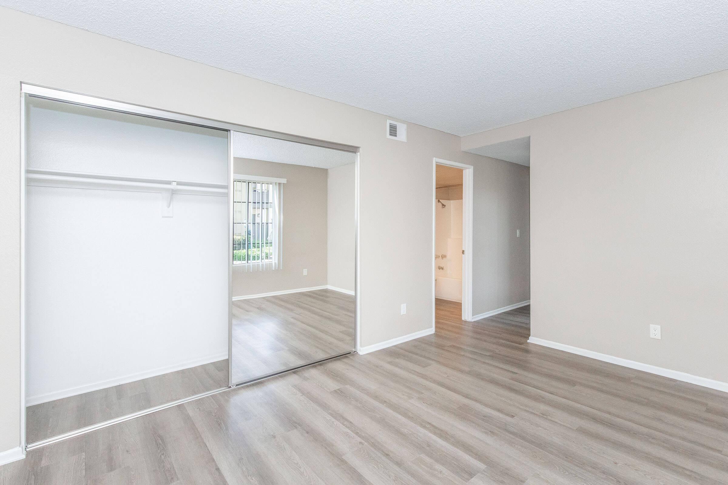 A spacious room with light-colored walls and hardwood flooring. It features a mirrored closet with sliding doors on the left and an open doorway leading to another area. Natural light filters through a window. The overall ambiance is bright and inviting, ideal for a living space.