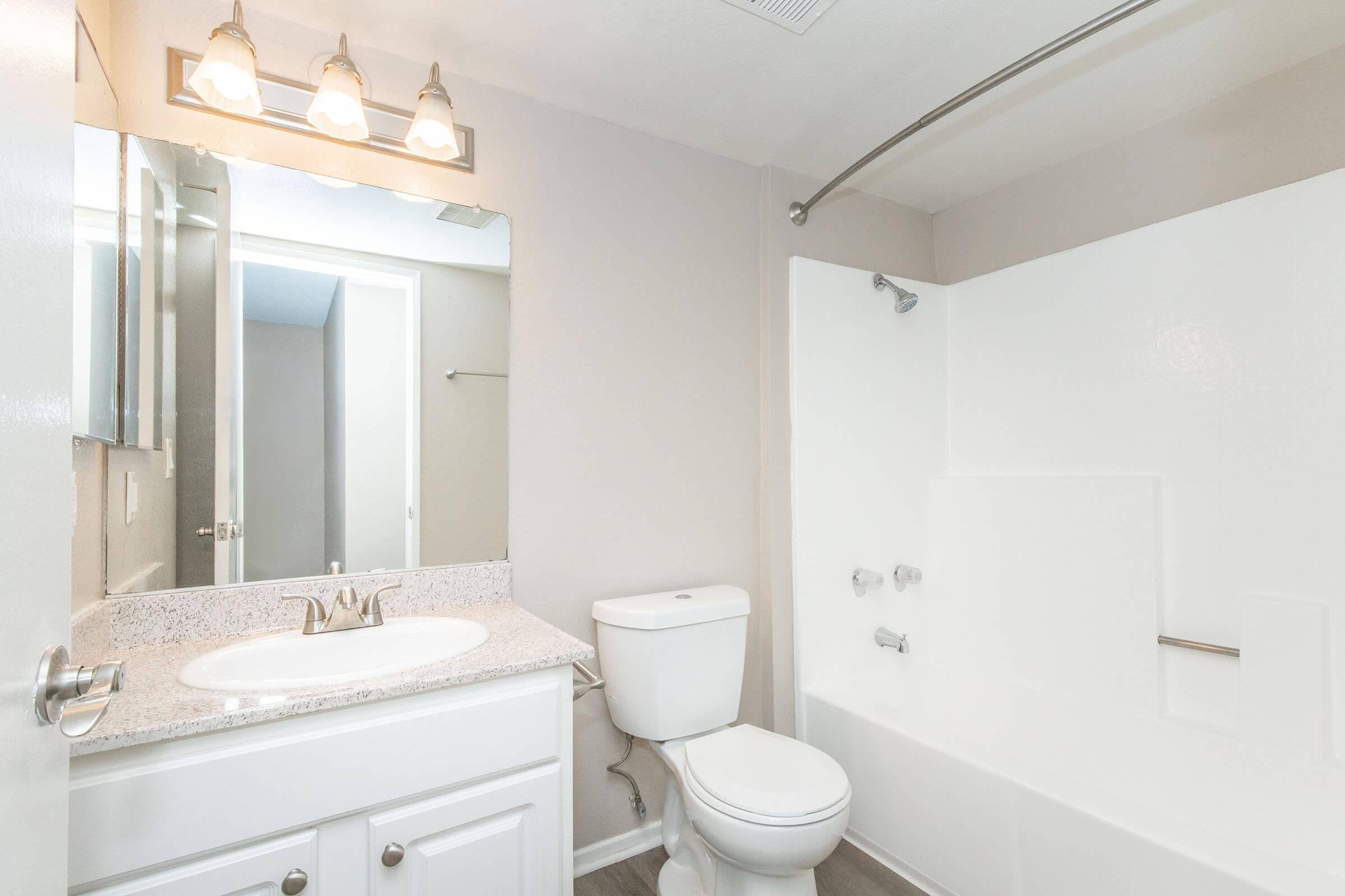 A modern bathroom featuring a white bathtub with shower, a toilet, and a sink with a granite countertop. There are three light fixtures above the sink, and a large mirror reflecting the space. The walls are painted in a light color, and the flooring is a wood-like laminate.