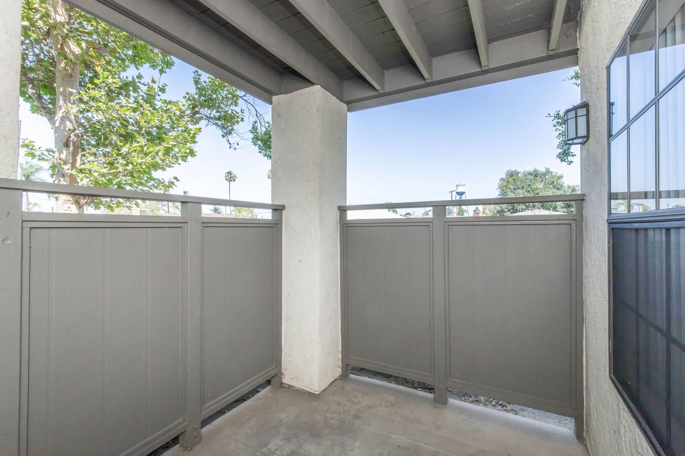 View of a small balcony or patio area with grey fencing, surrounded by greenery. The space is well-lit, featuring a concrete floor and an overhang supported by beams. A window is visible on one side, contributing to the open atmosphere.