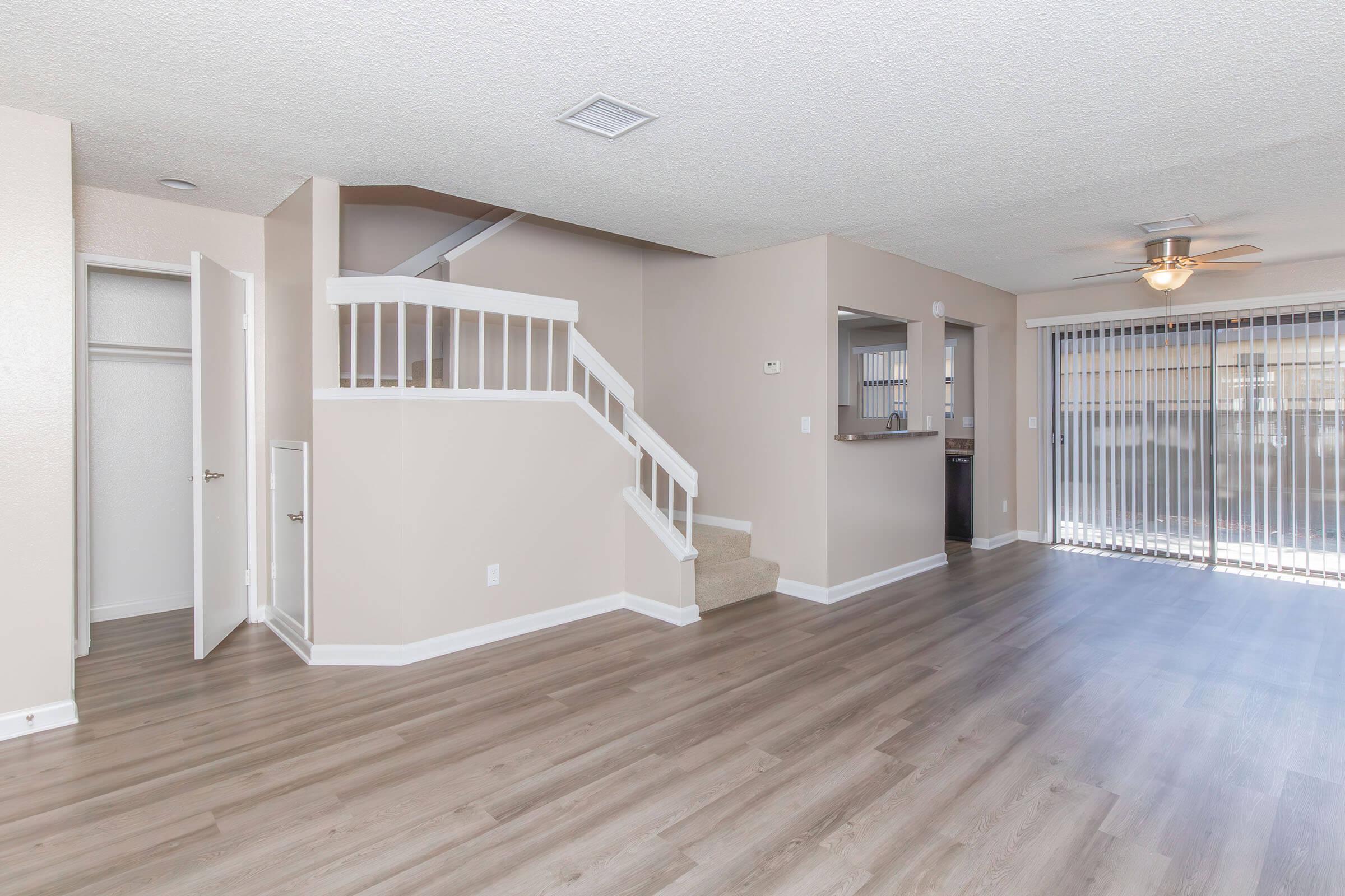 Interior view of a spacious living area featuring light-colored walls, hardwood flooring, and a ceiling fan. Stairs lead to an upper level, while a door opens to a private outdoor space with sliding glass doors. The room is well-lit, creating a welcoming atmosphere.