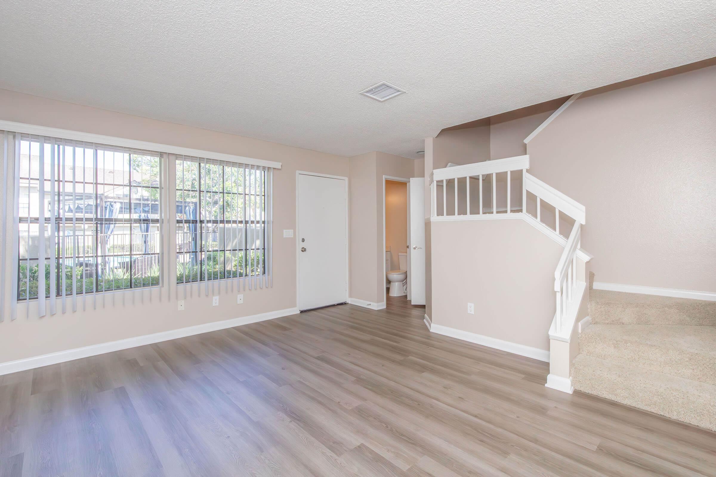 A spacious, well-lit living room with large windows featuring vertical blinds, light-colored walls, and new laminate flooring. A staircase with white railings leads to an upper level. A door on the left leads outside, and a small bathroom is visible through an open door at the rear.