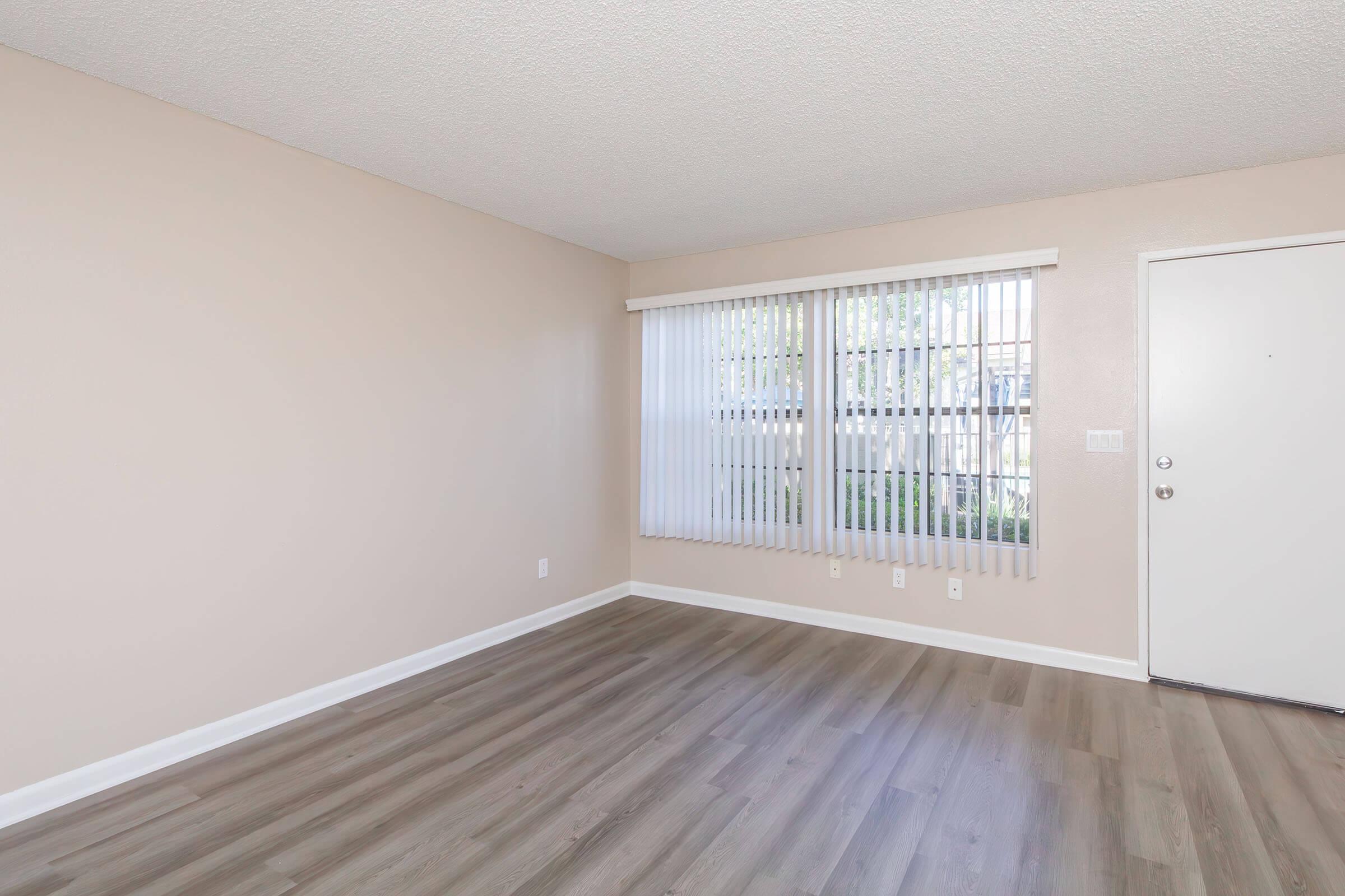 Empty room with light-colored walls and laminate flooring. Large window with vertical blinds allows natural light to enter, and a door is visible on the right side. The room appears spacious and well-lit, suitable for various uses.