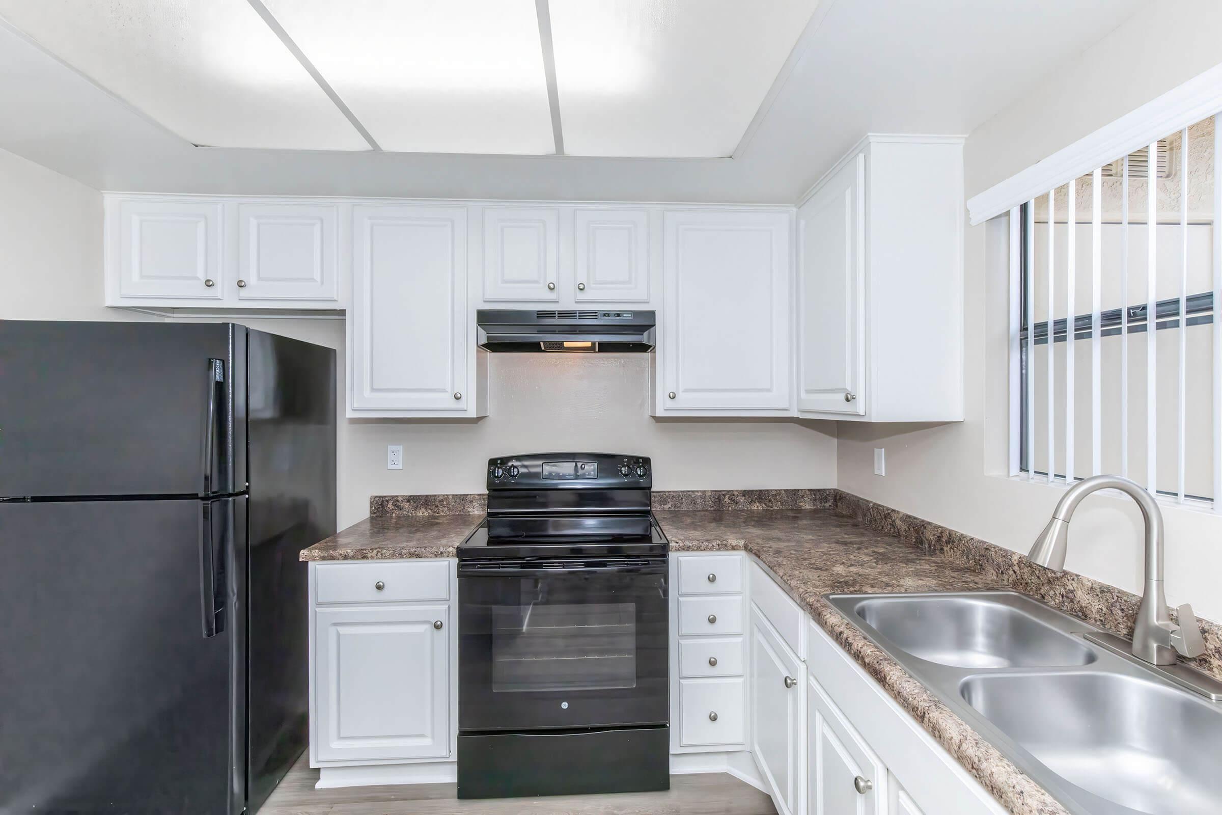 Modern kitchen featuring white cabinetry, a black refrigerator, and a black stove with an overhead exhaust fan. The countertop is a textured dark color, and there are dual sinks. Natural light filters in through horizontal blinds on the window.