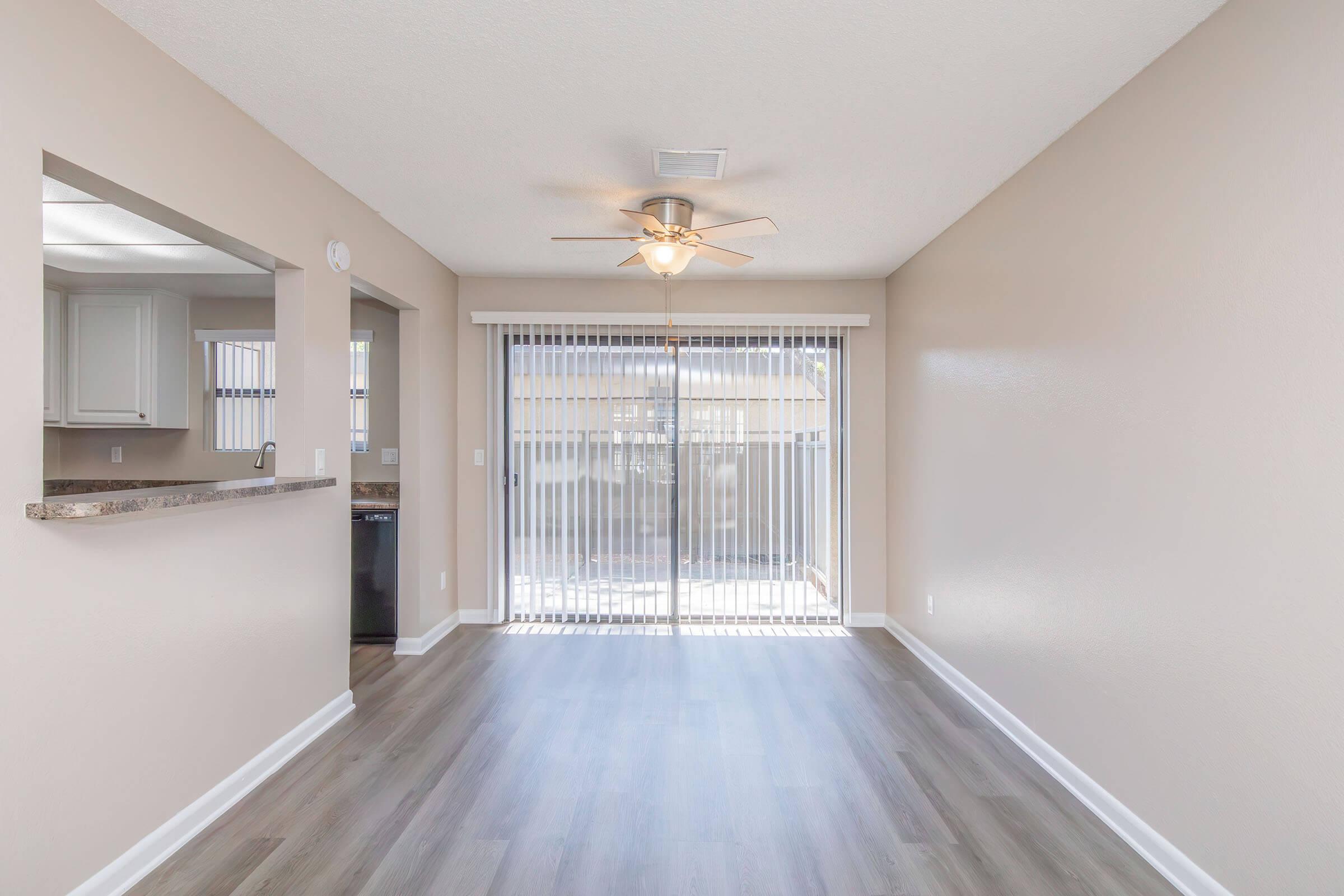 Bright and airy living room with light-colored walls and a ceiling fan, featuring large sliding glass doors that lead to an outdoor space. A small open area with a bar overlooks a kitchen area, showcasing modern flooring and a spacious layout. Natural light fills the room, enhancing the inviting atmosphere.