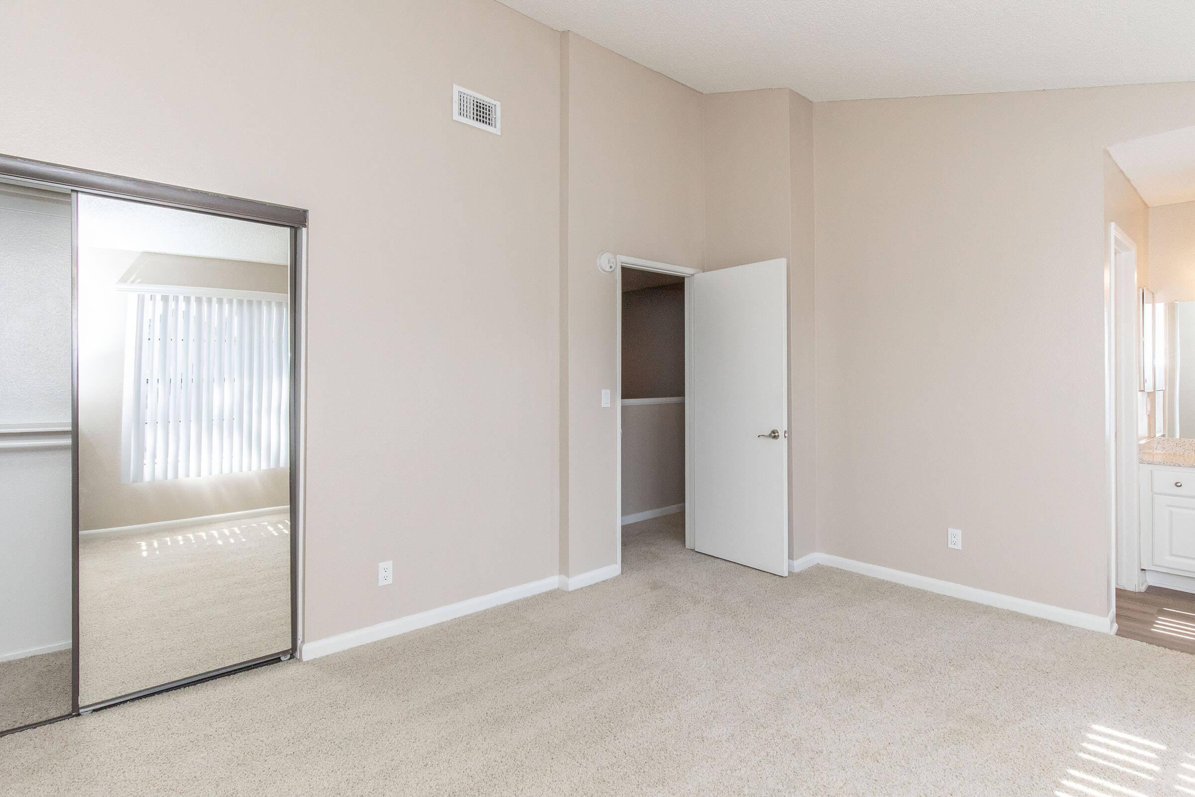 A well-lit, neutral-colored bedroom featuring plush carpet, a large mirrored closet on one side, and an open doorway leading to another area. The walls are painted a soft beige, and there are vertical blinds on a nearby window, allowing natural light to fill the space.