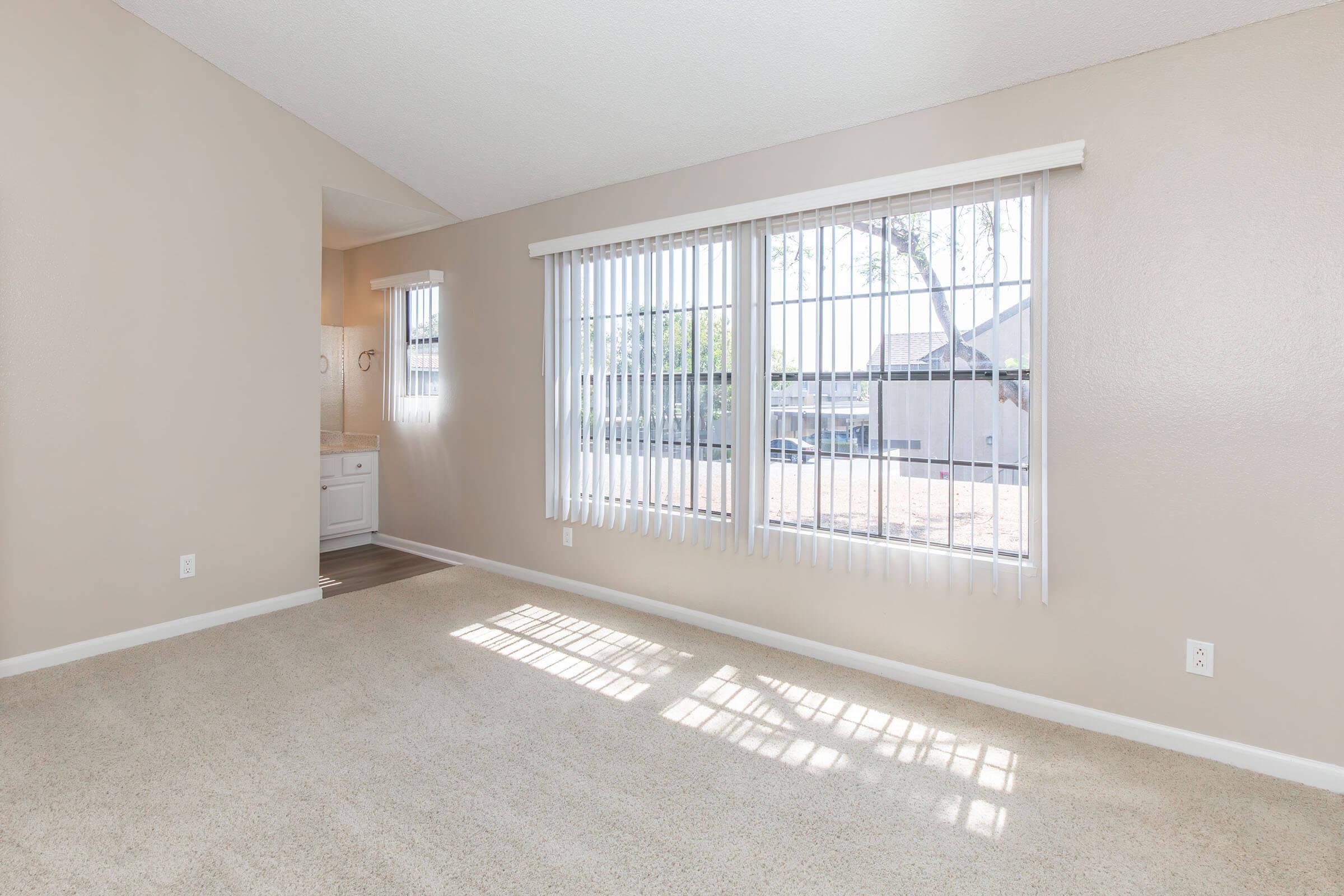 A sunlit room with beige walls and a carpeted floor, featuring large windows with vertical blinds. The windows allow natural light to stream in, casting shadows on the floor. In the background, a glimpse of a kitchen area is visible through an open doorway.