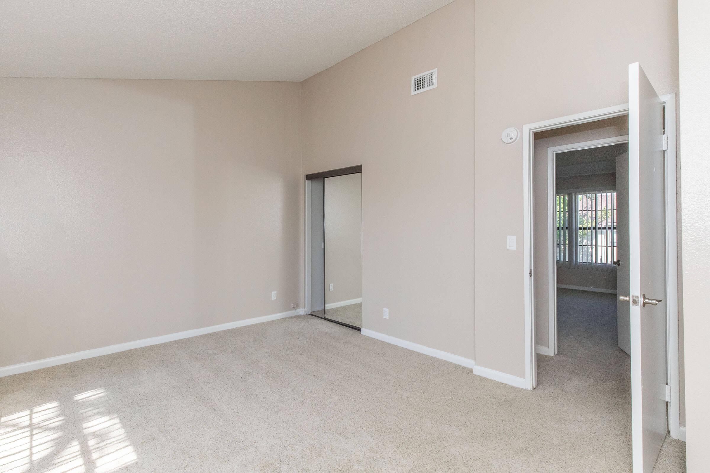 Spacious room with light-colored walls and plush carpeting. A door leads to another room, while a mirror occupies one wall. The room features a tall ceiling, providing an open feel. Natural light streams in through the windows, creating a bright and inviting atmosphere.